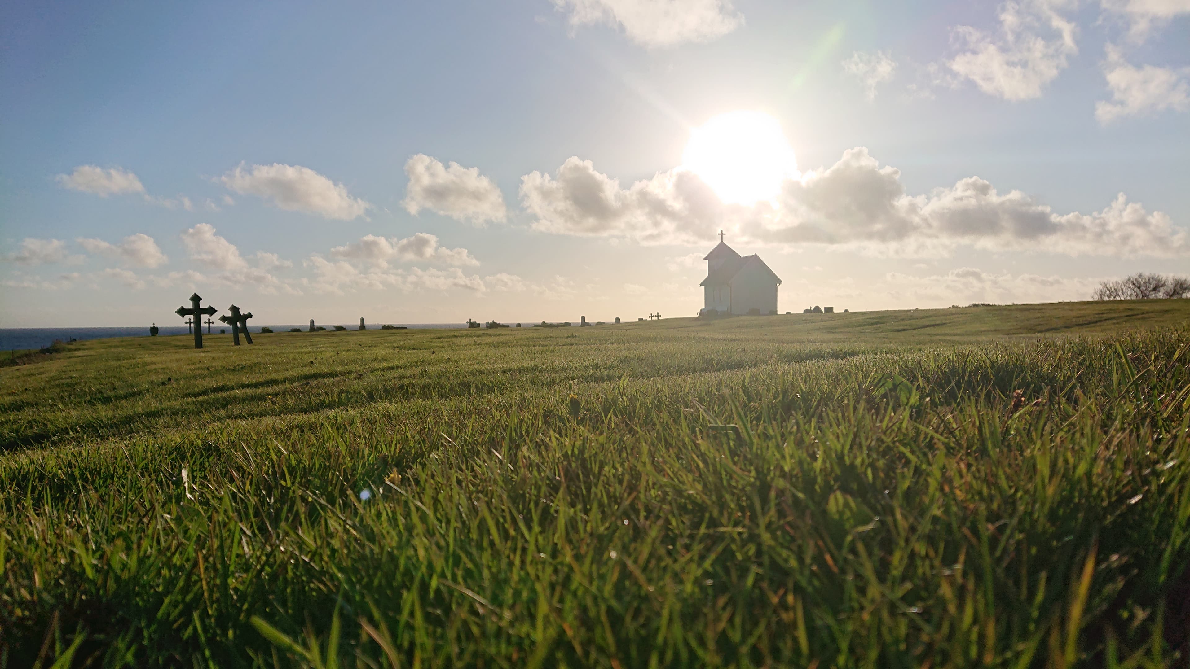 Varhaug gamle kirkegård i det fjerne med grønne beitemarker foran samt noen steinkors. Solen titter frem fra noen lette skyer på himmelen.