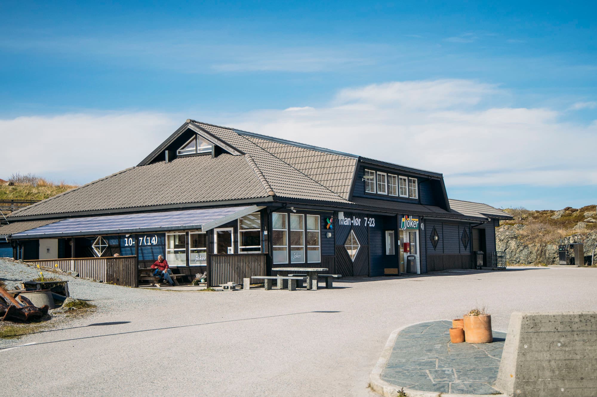 joker butikk i trebbygning. person sitter utenfor. parkering utenfor butikken