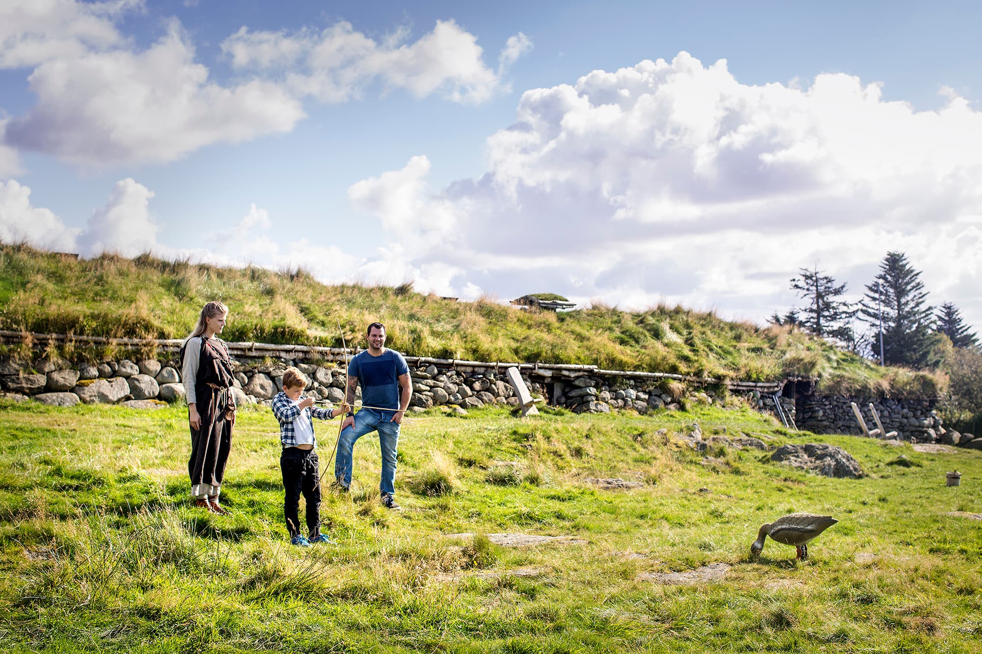 Et barn skyter med pil og bue utenfor langhuset på Jernaldergården. To voksne ser på.