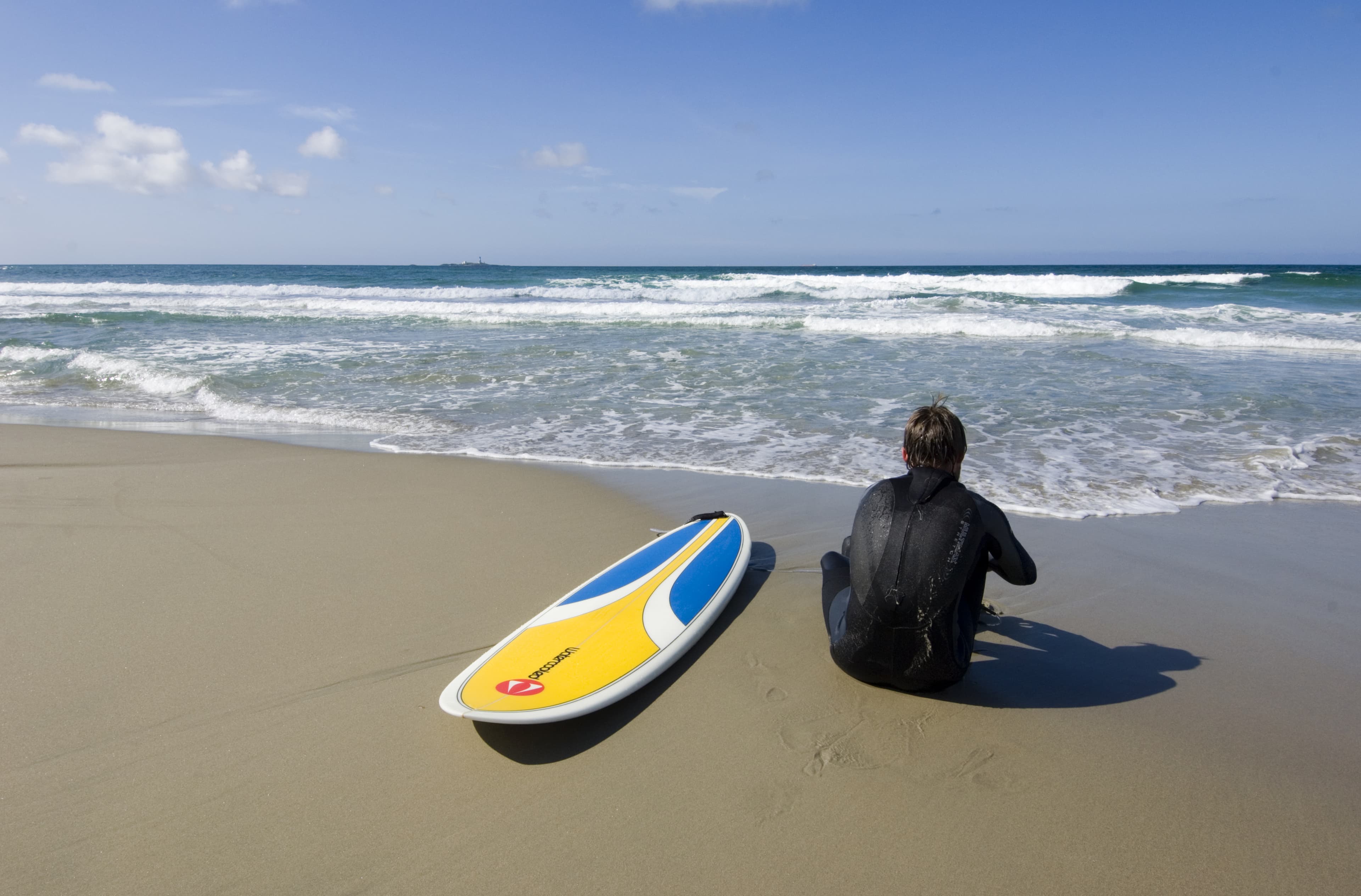 En surfer sitter i sanden med surfebrettet ved siden av seg og ser ut mot bølgene.