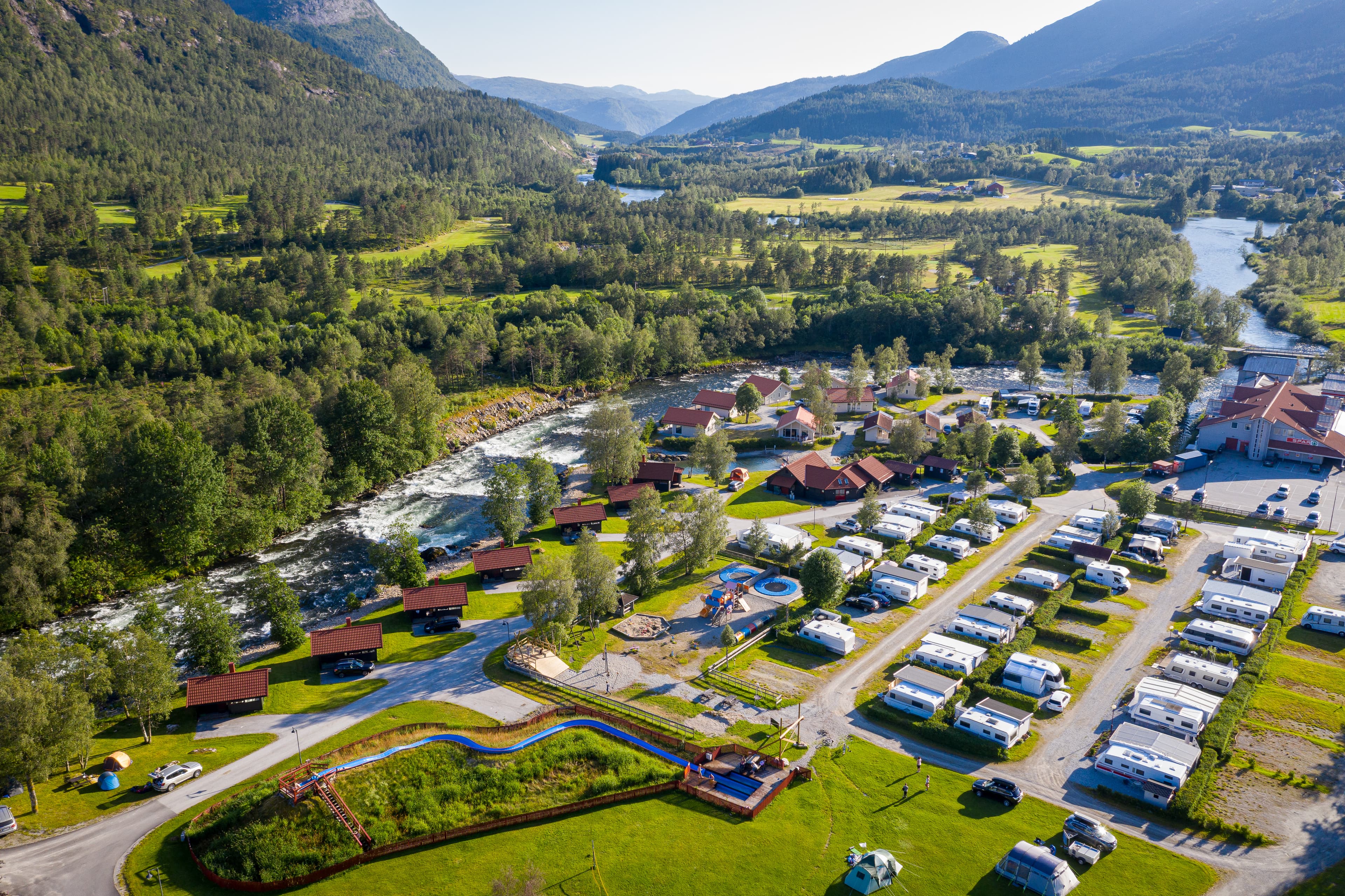 Dronebilde. Vi ser rett ned på bubil og campingvognparkeringa, med teltplassar og leieplass. Langs elva ligg hyttene. Til høgre i bildet ligg servicehuset, butikk, bensinstasjon og serveringstad. Vi ser elva renne nedover dalen. Det er mykje frodig skog og vi ser starten på nokre av fjella.