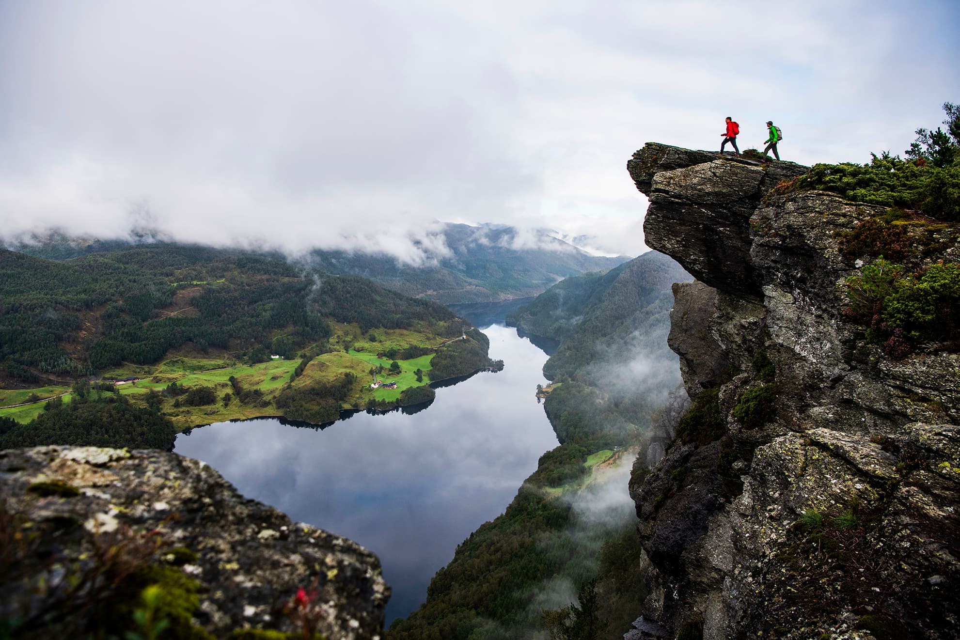 To personer som står på kanten av et fjell og ser utover landskapet og vannet nedenfor.