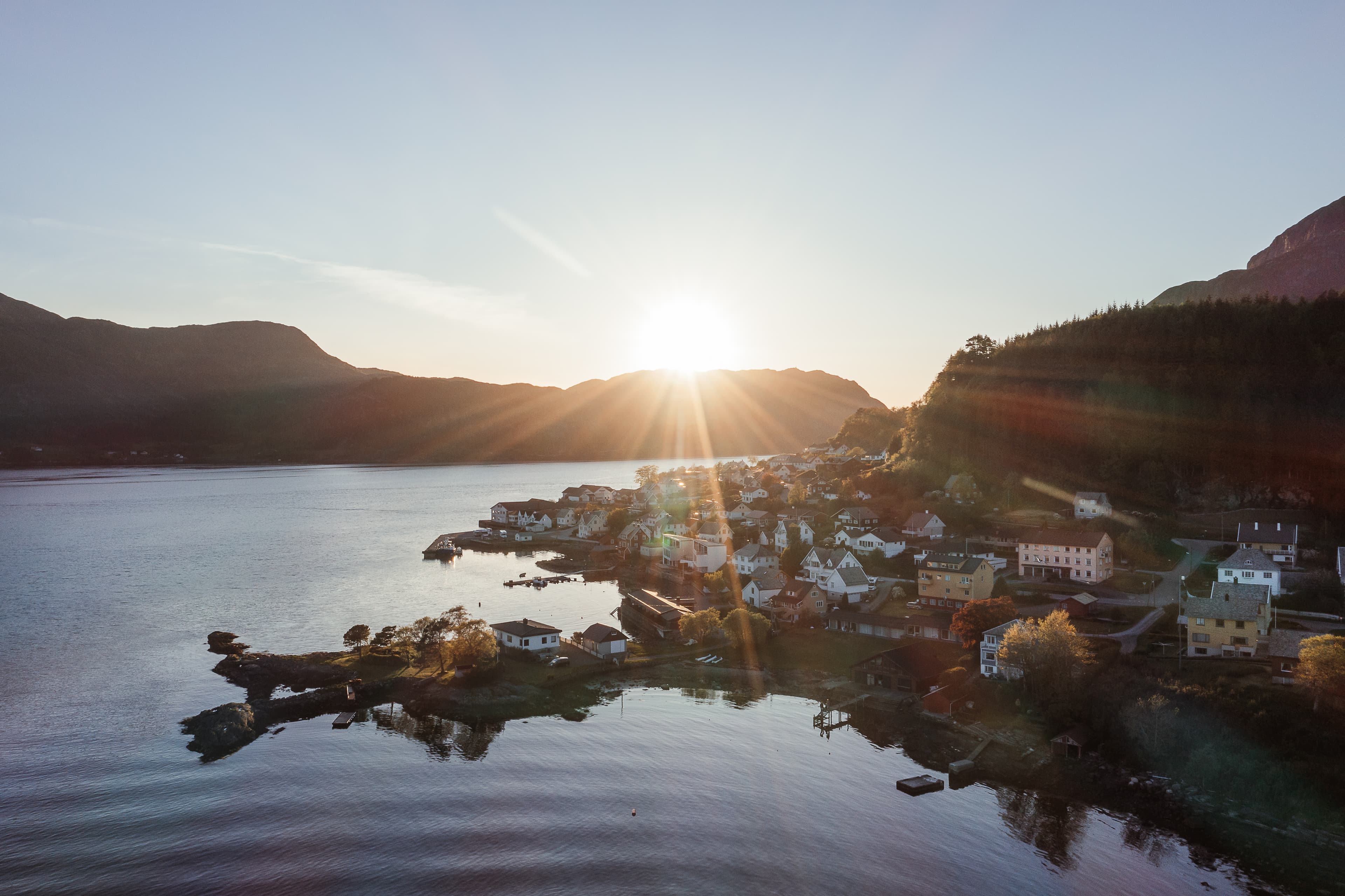 Vi ser bebyggelse som ligg ved sjøen. Sola held på å gå ned bak fjellet. Det er blå himmel. Foto