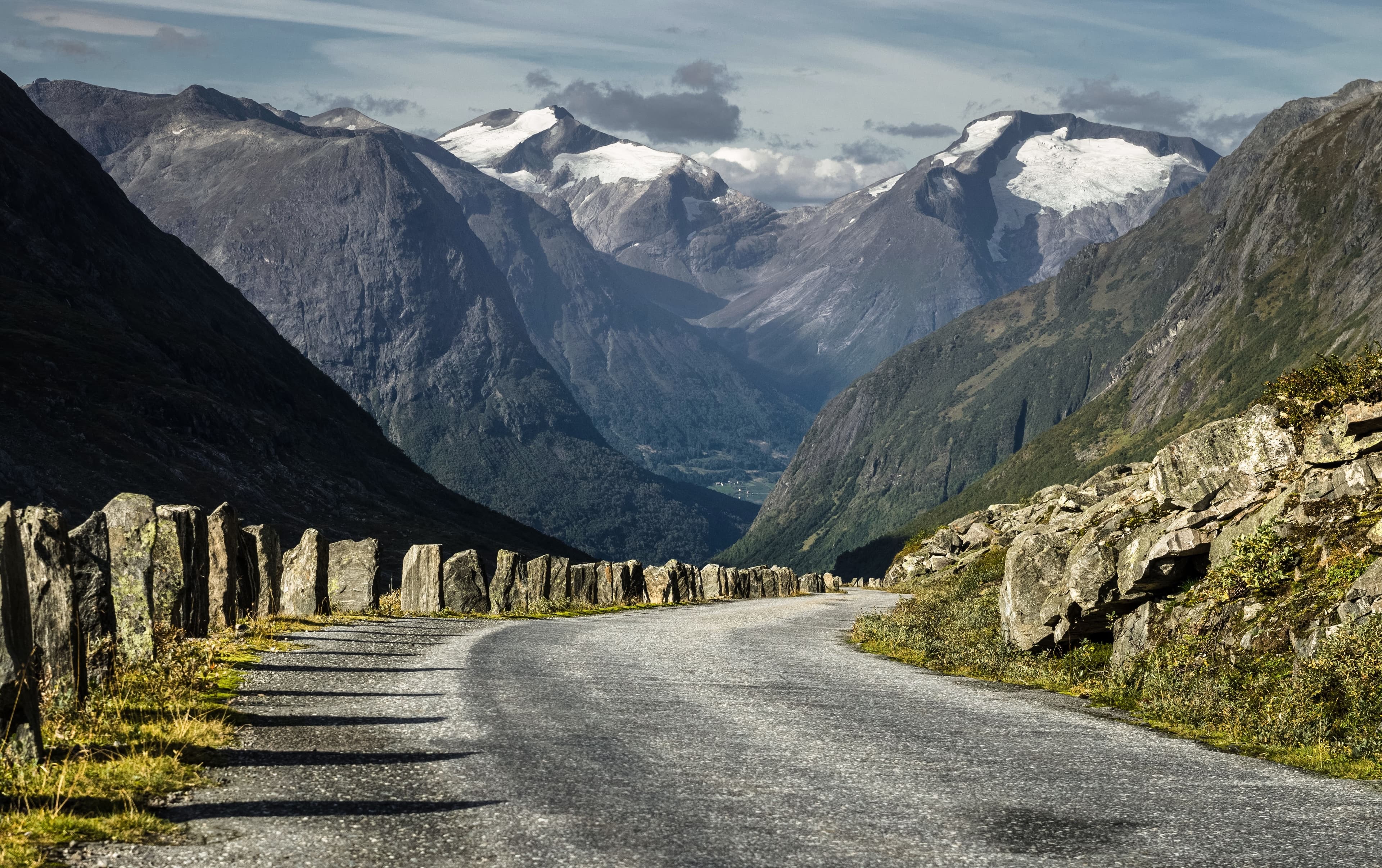 En vei som går nedover en dal med stabbesteiner og mektige, delvis snødekkede fjell i bakgrunnen.