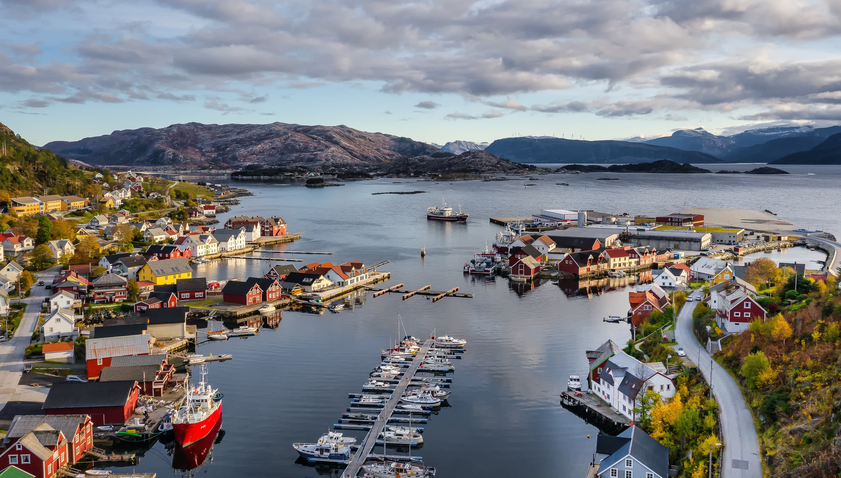 En havn med flere båter og hus rundt havnen. Øyer og fjell i bakgrunnen.