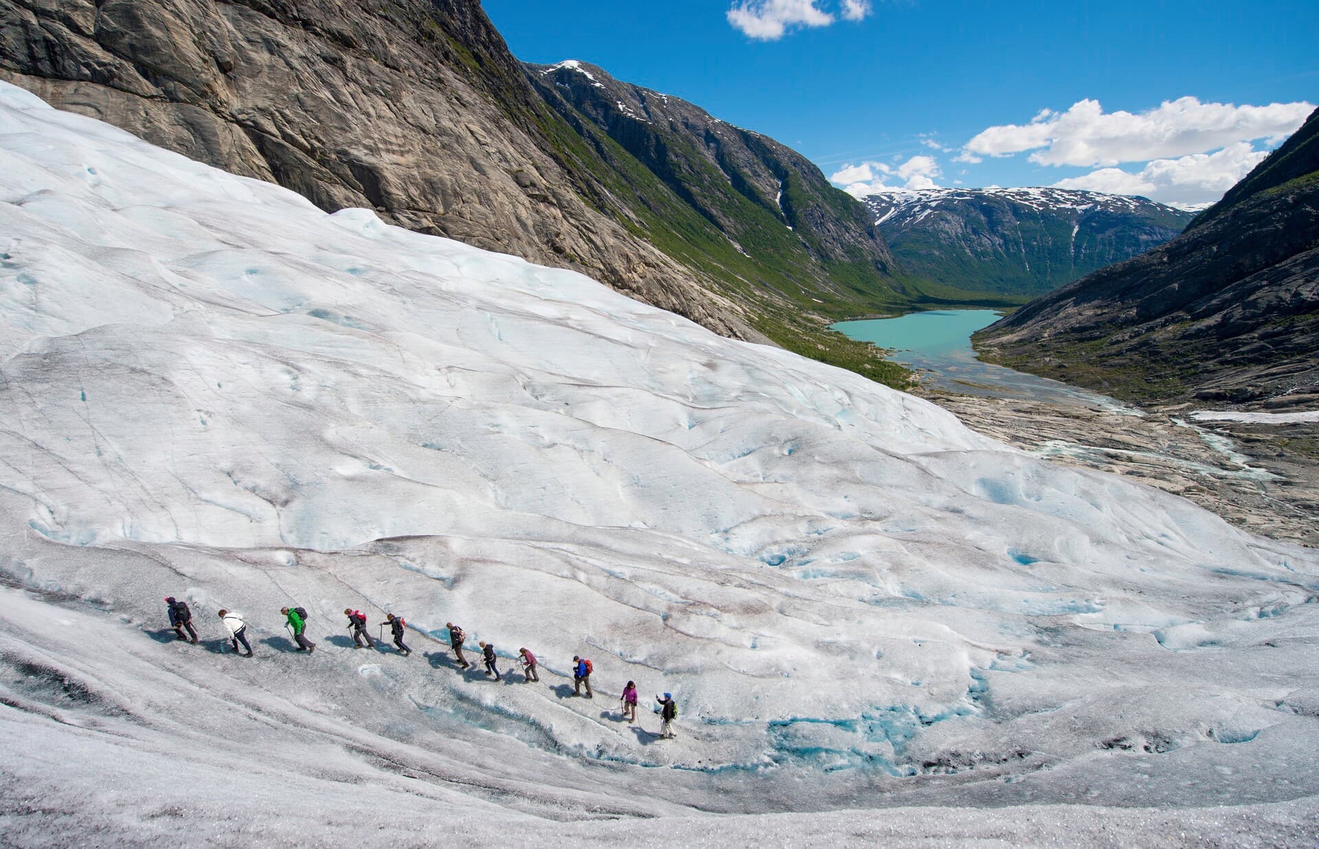 En gruppe mennesker som går i tau på en isbre, med en innsjø lenger nede.