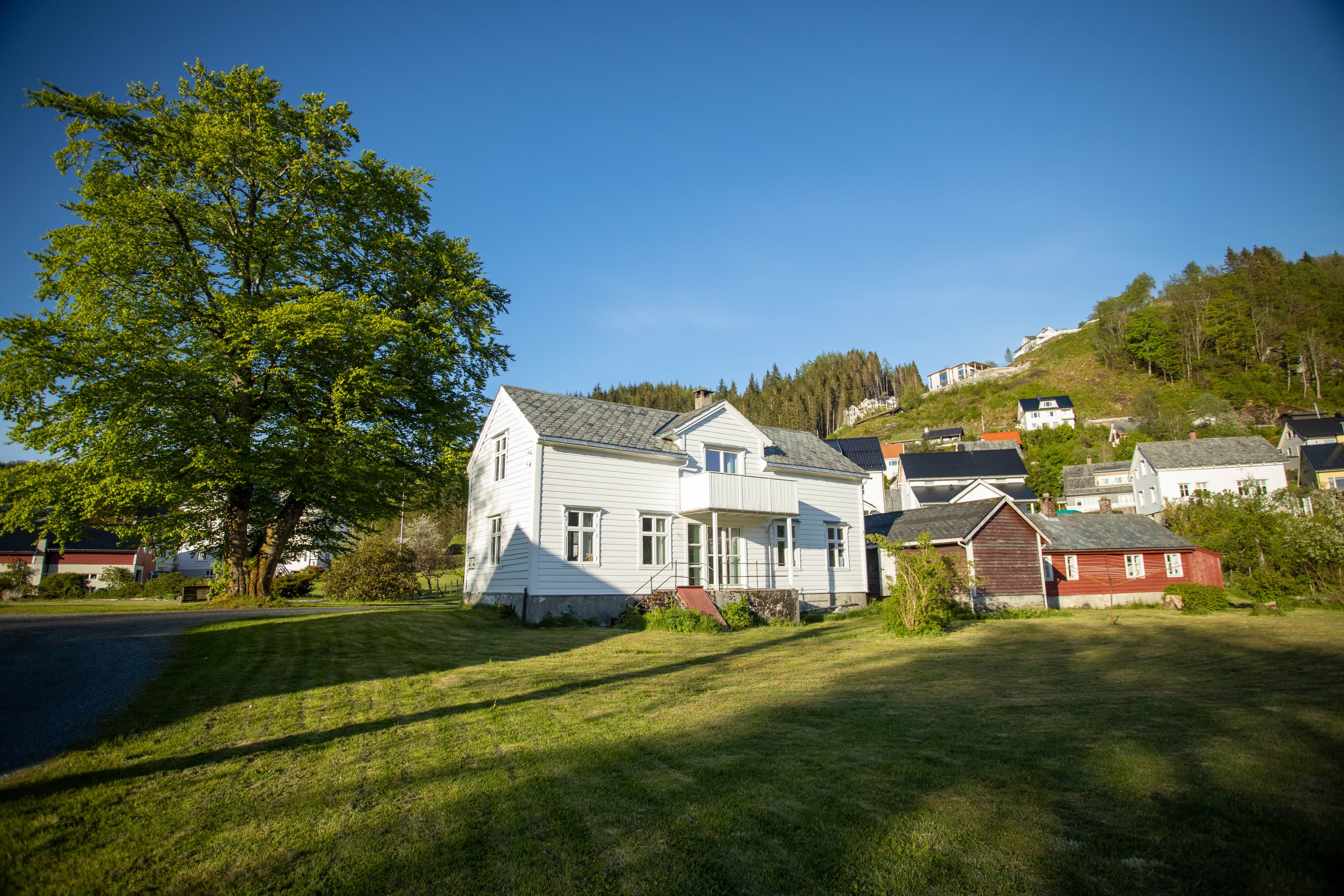Jakob Sande-tunet. Det kvite hovedhuset med nokre små raude hus. Til venstre for hovedhuset står eit stort grønt tre. Bak huset ser vi meir bebyggelse. Framfor huset er det stor plen. Det er sol og blå himmel. Foto