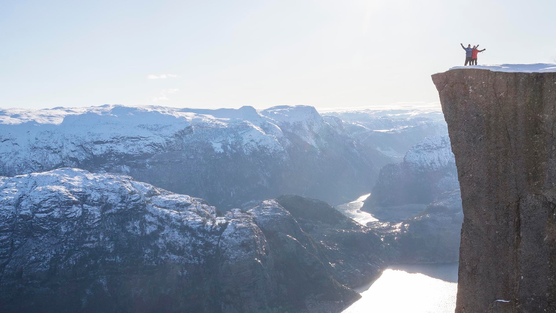 Tre personer som står på Preikestolen med snødekte fjell i bakgrunnen.