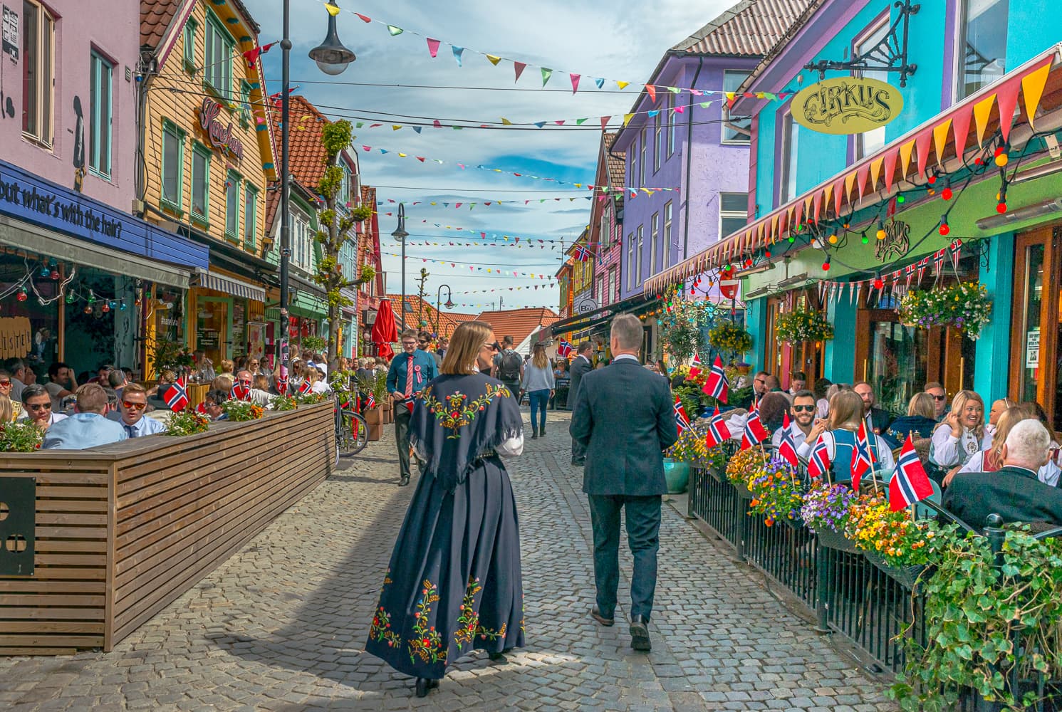 Par går nedover Fargegaten på 17. mai i norsk bunad. Gaten er pyntet med norske flagg.
