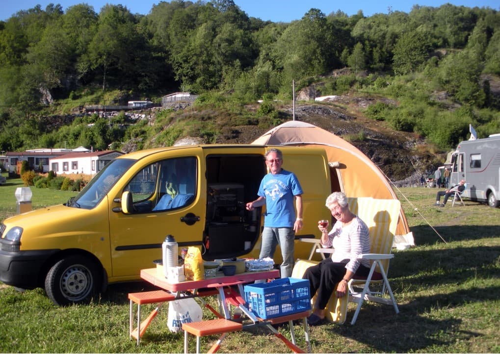 To campinggjester smiler til kamera mens de spiser ved campingbord foran teltet og bilen sin.
