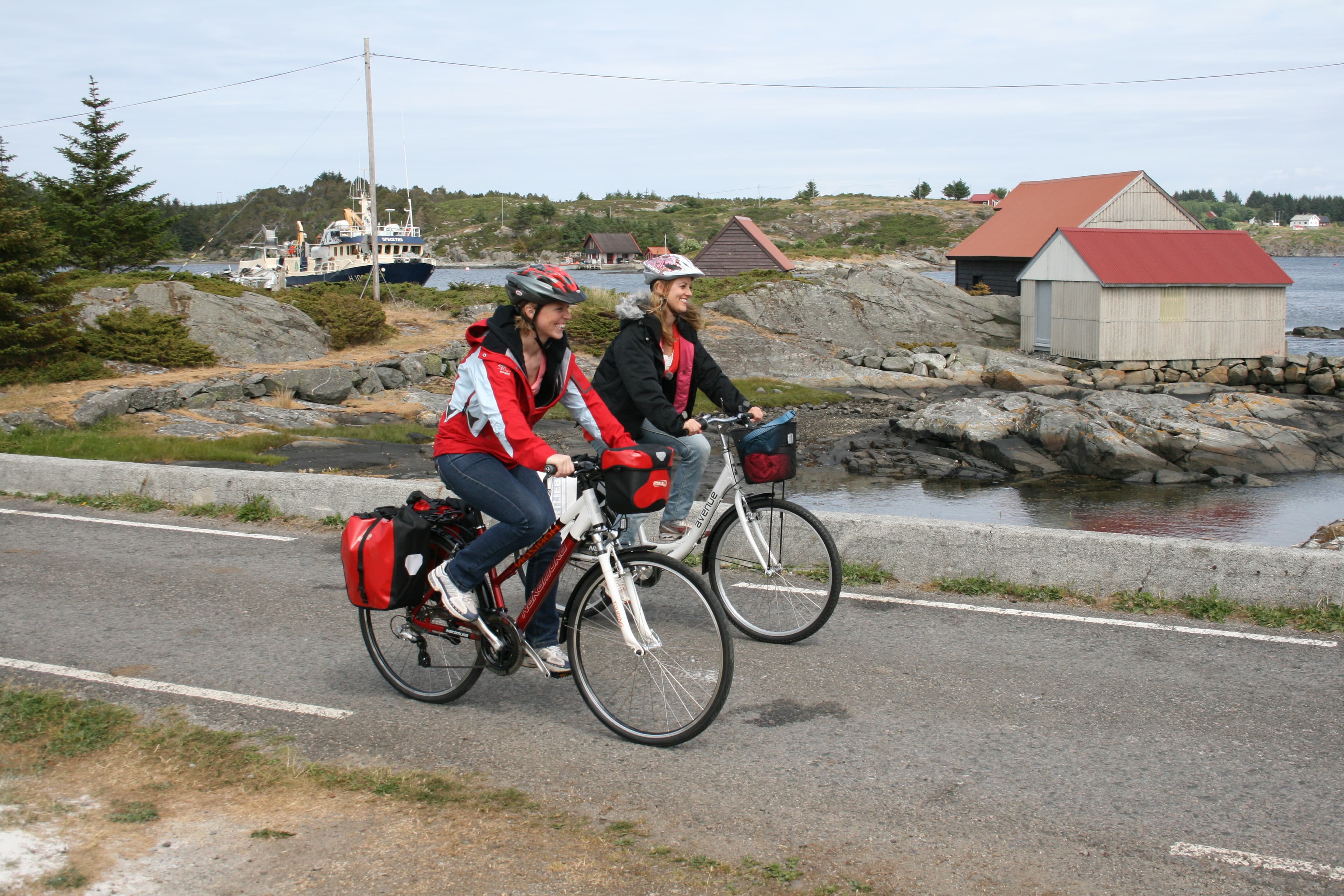 To personer som sykler ved sjø, båt og naust.