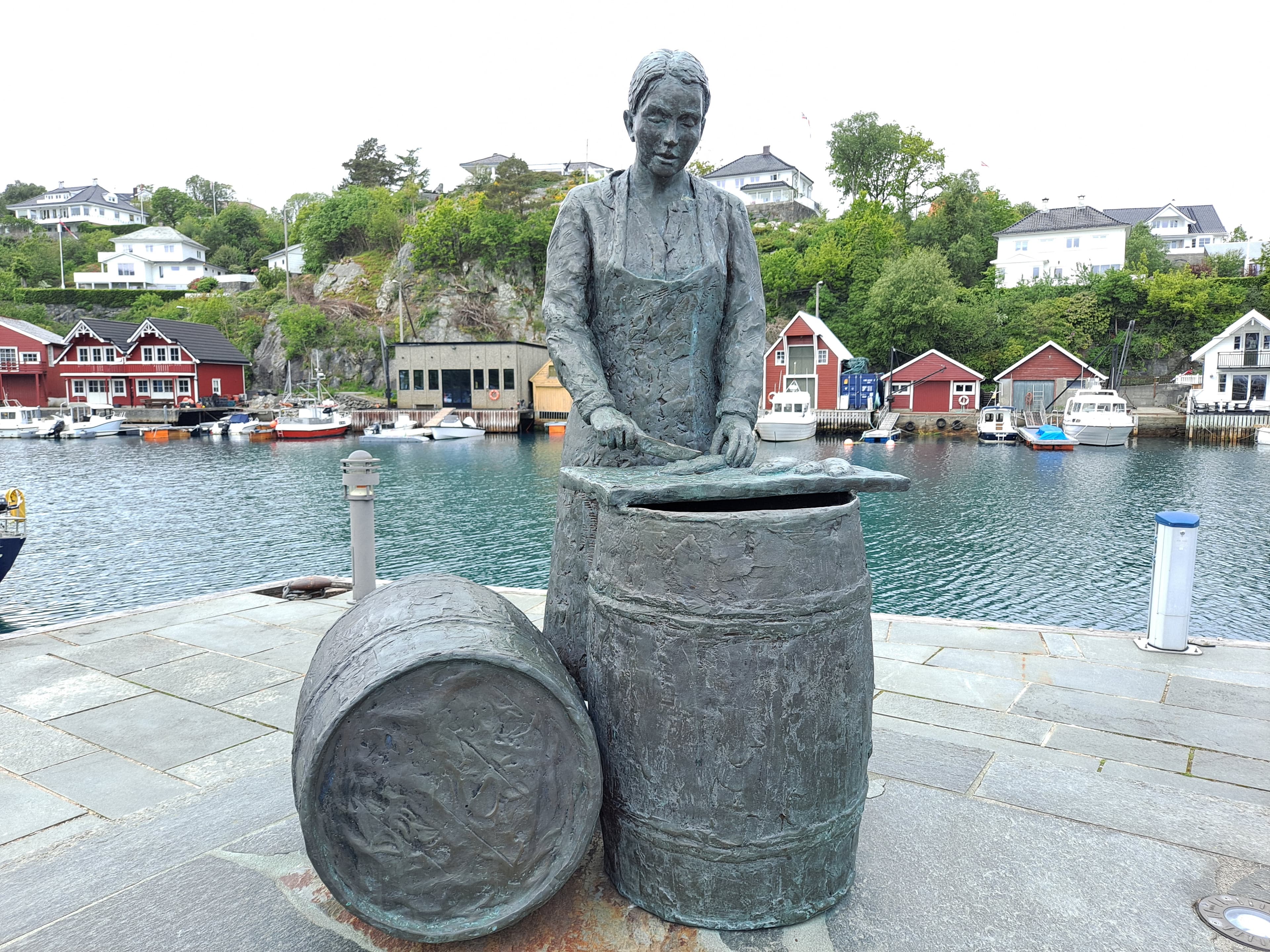 Statue av jente som rensker sild på tønnefat ved sjø.