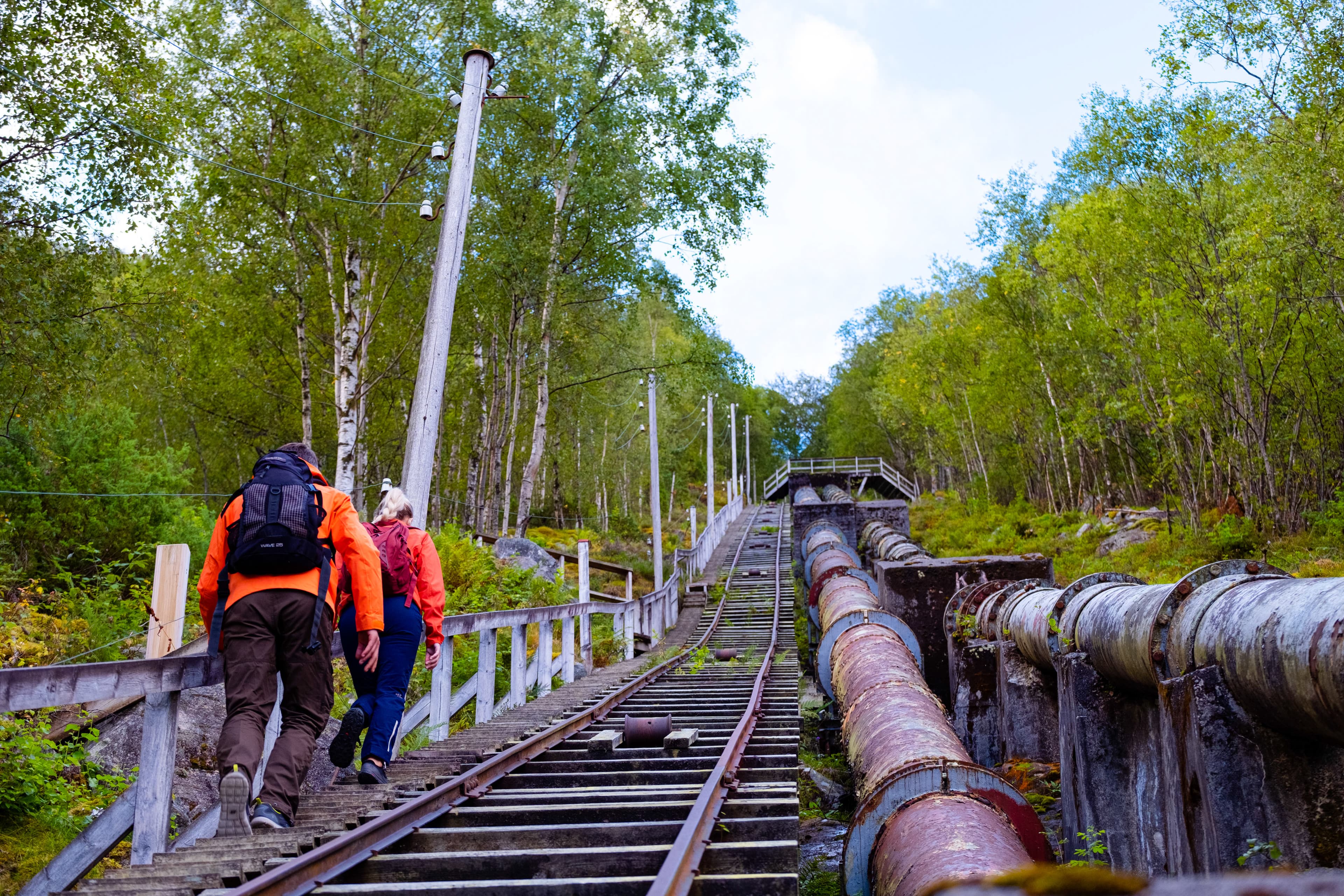 Et par med oransje jakker på vei opp tretrappene Flørli. Grønne trær på hver side. Bratt fjellside.