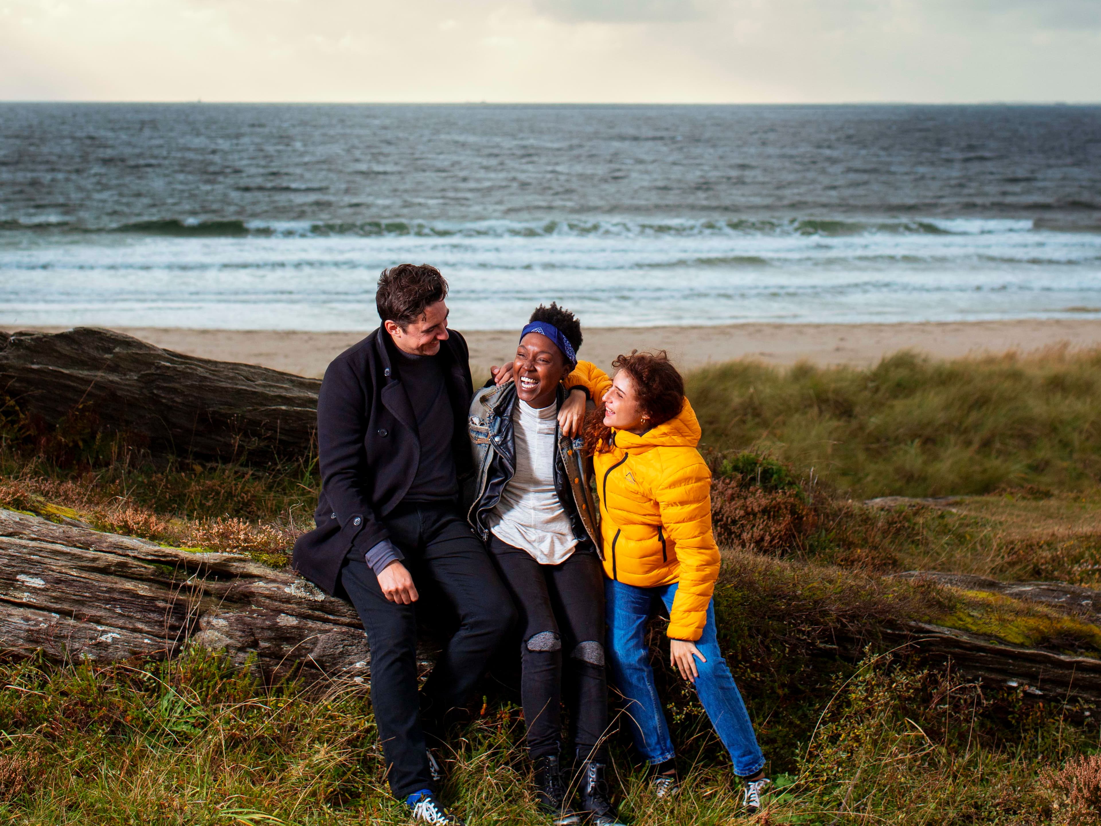 Tre personer sitter på et svaberg på Solastranden med stranden bak seg. Himmelen er litt overskyet med solen som gløtter frem mellom skyene. Høstfarger på bakken.