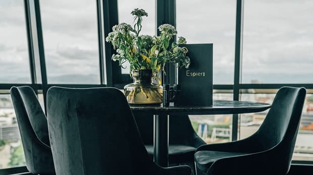 Rundt bord med fire stoler med barmeny på bordet. En vase med friske blomster på bordet. Fin utsikt mot by.