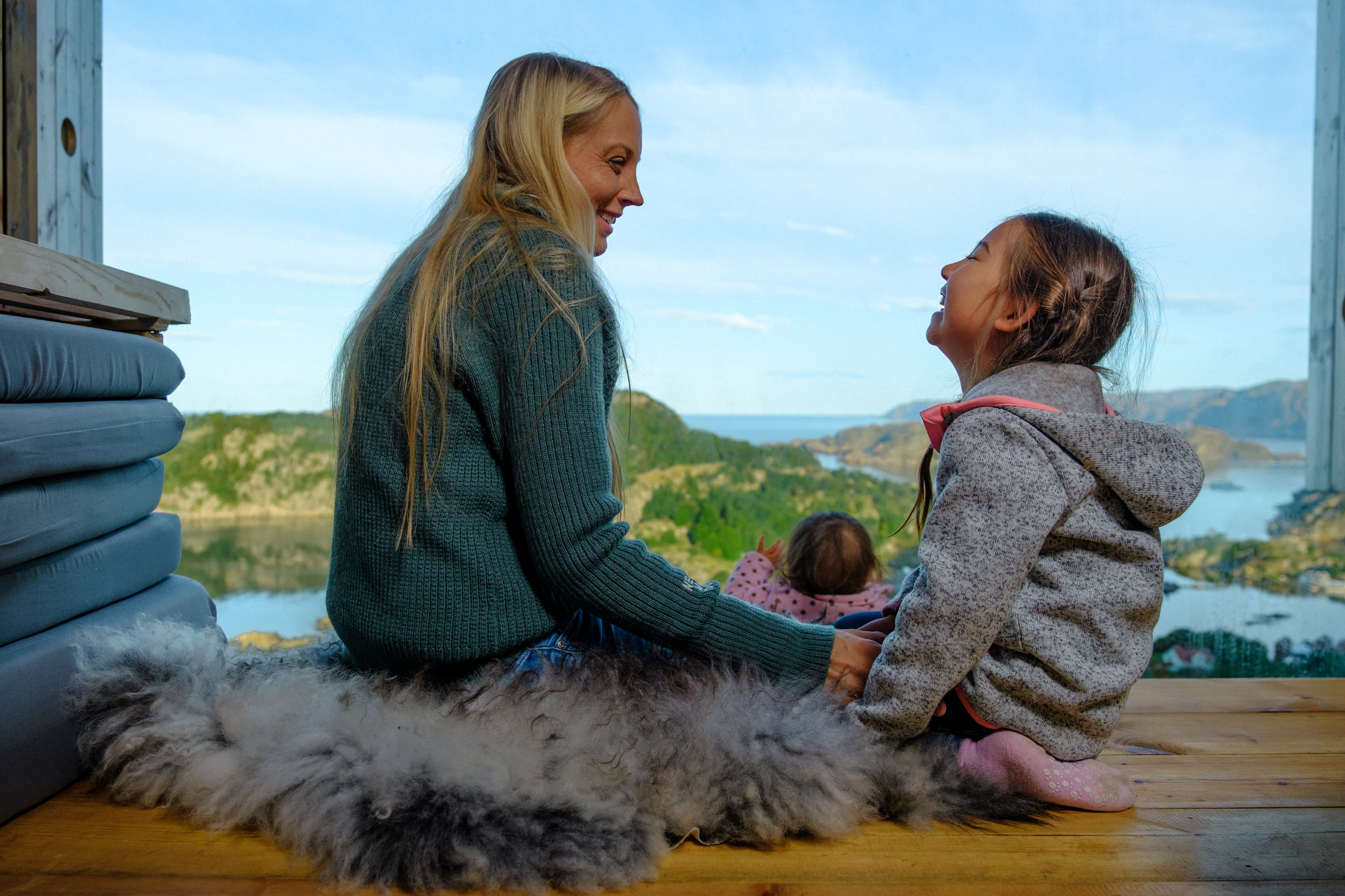 Mor med to barn sitter på en skinnfell og ser utover fin natur.