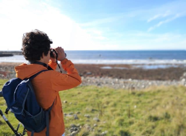 En gutt i oransje jakke og blå sekk som ser ut over havet med kikkert