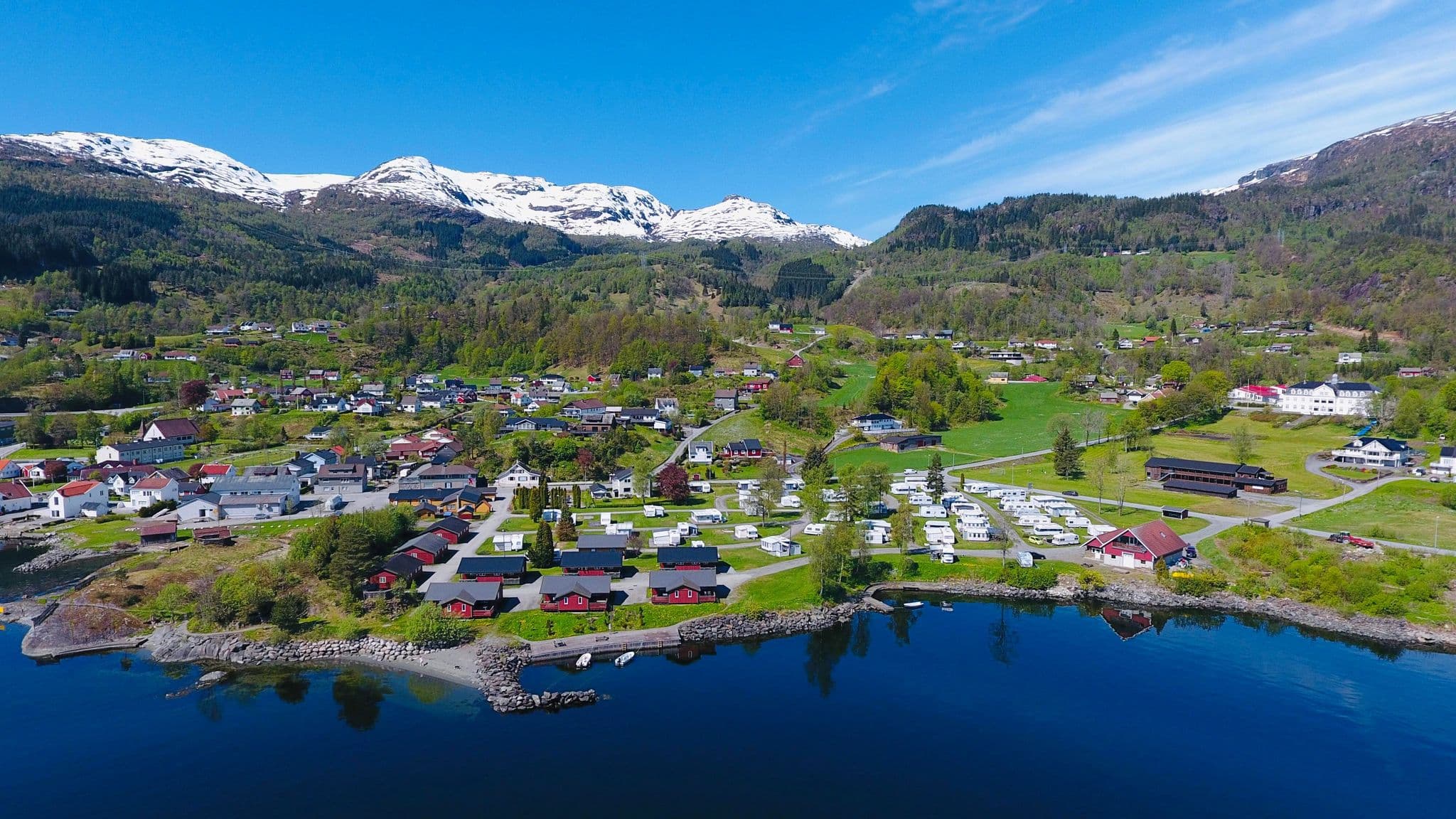 Oversiktsbilde av campingplassen med fjorden i forgrunn og snøkledde fjell i bakgrunnen.