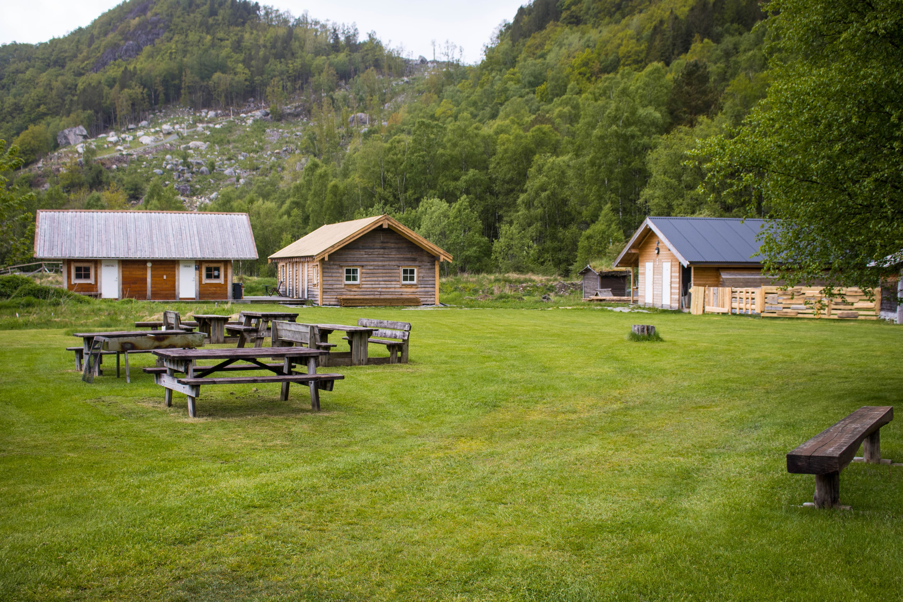 To hytter og sanitæranlegget i bakgrunnen og et grønt område med benker og bord i forgrunnen.