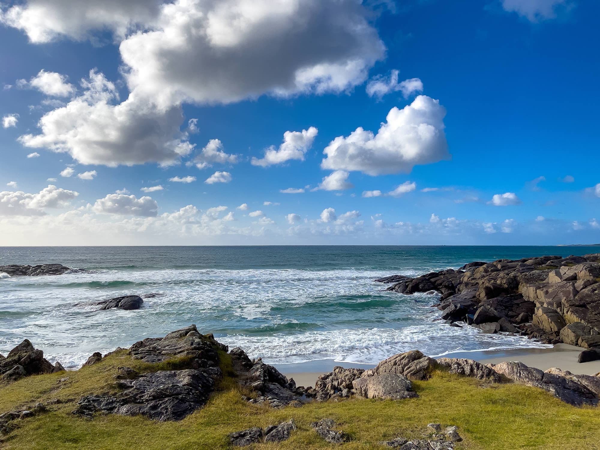 Holmasanden med blå himmel og hvite skydotter. Havet bruser inn mot land med svaberg og strand.