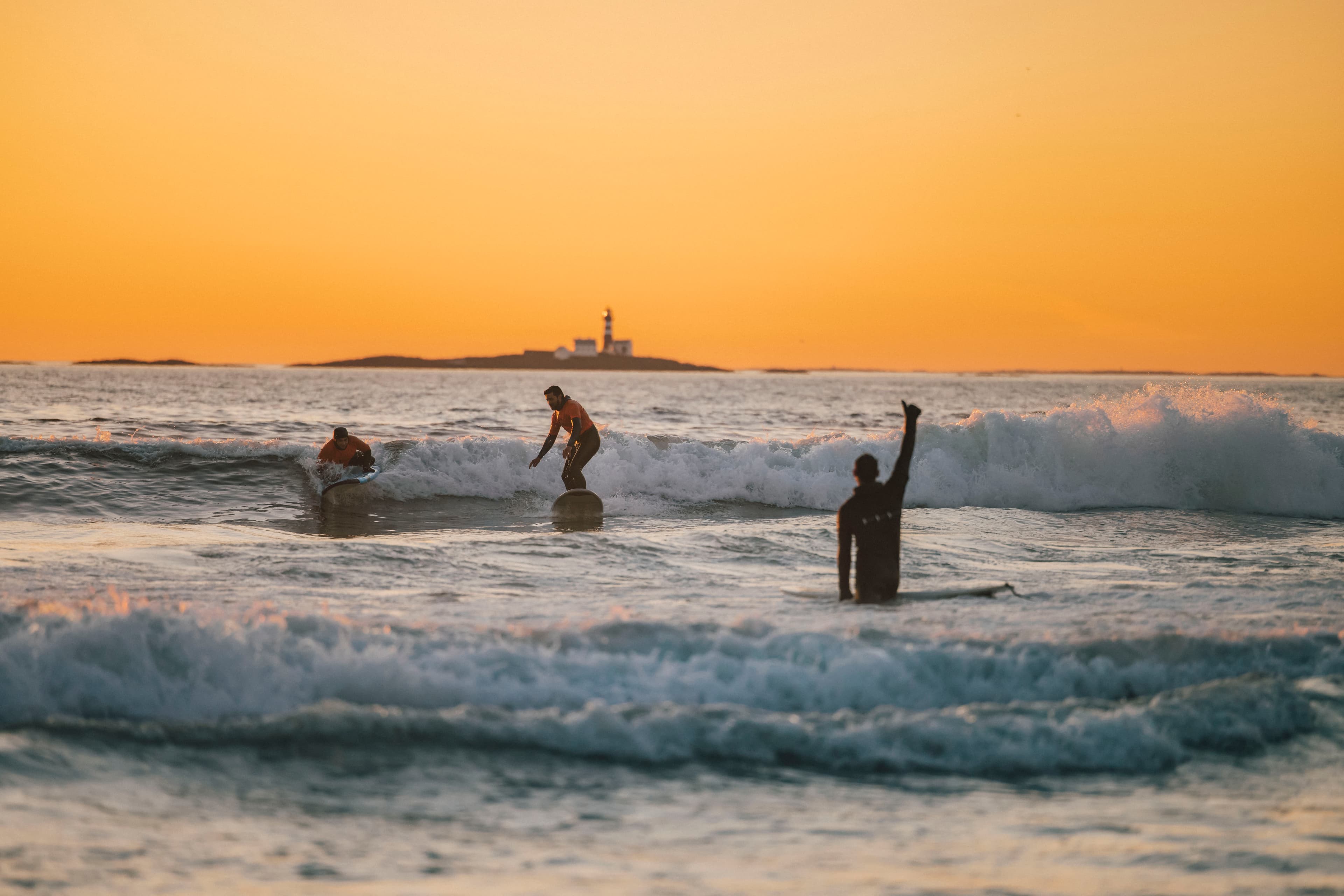 Three people in the water with surf boards in a yellow evening sun. The lighthouse Feistein in the distance