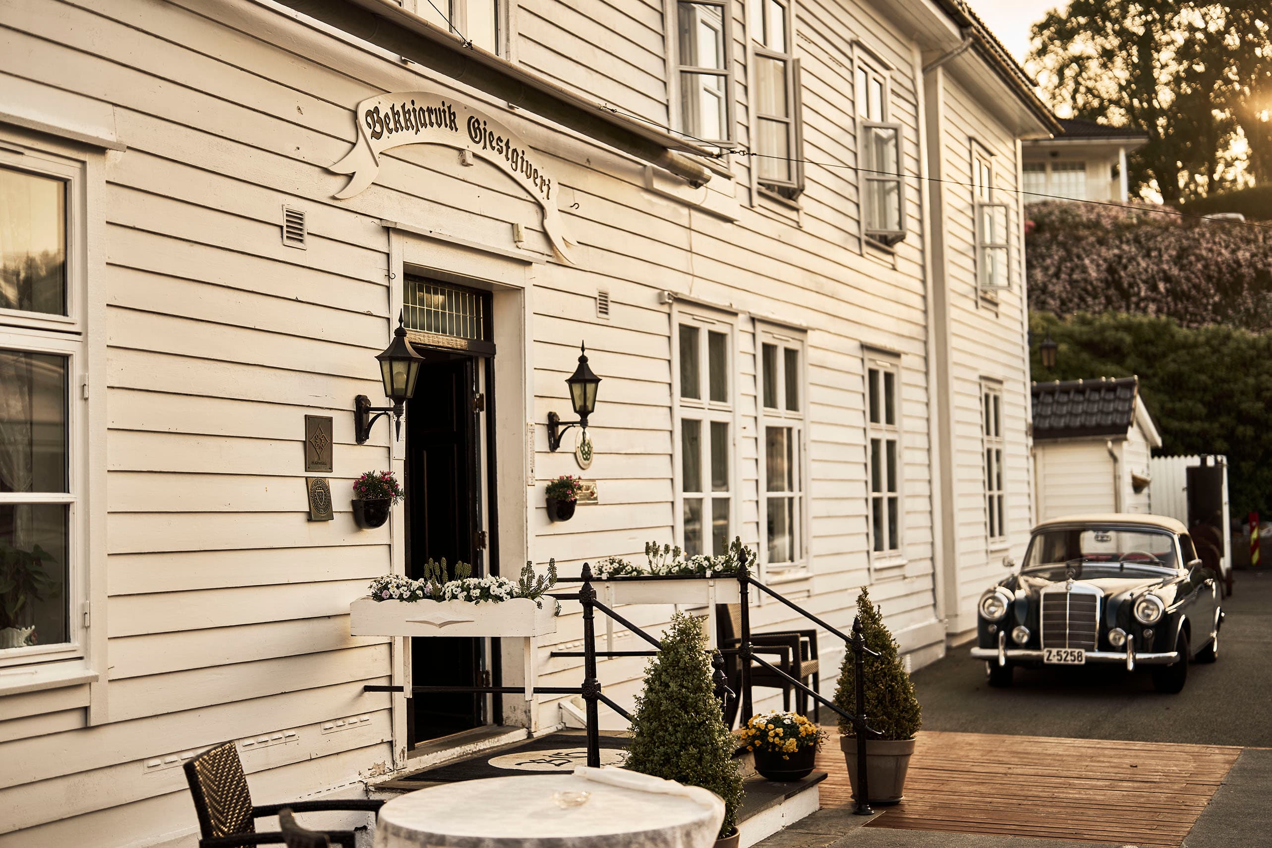 Fasade på gjestgiveri i hvitmalt tre,  sort veteranbil parkert ved veggen