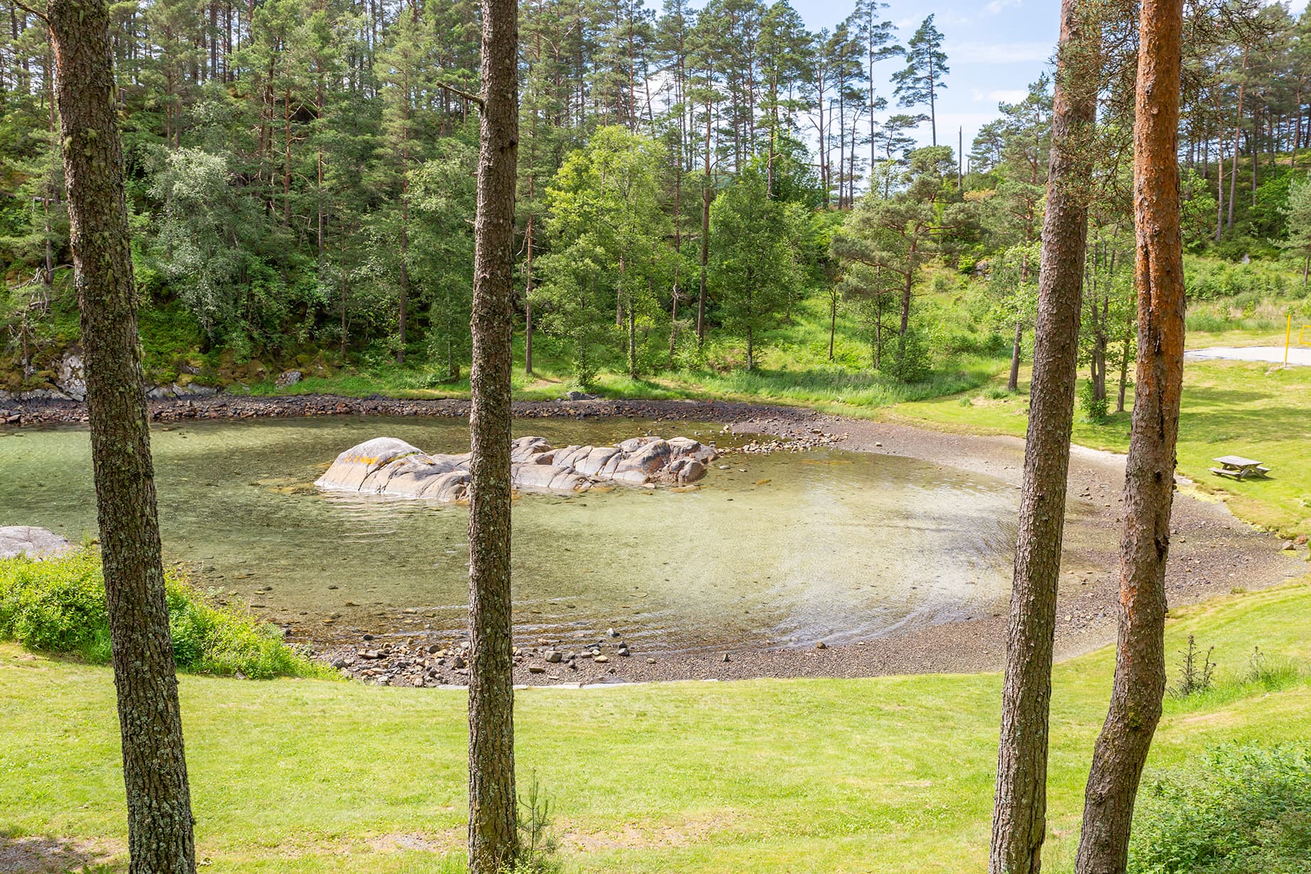 Badestrand omgitt av grønne bakker, trær, en benk med bord til står på en plen.