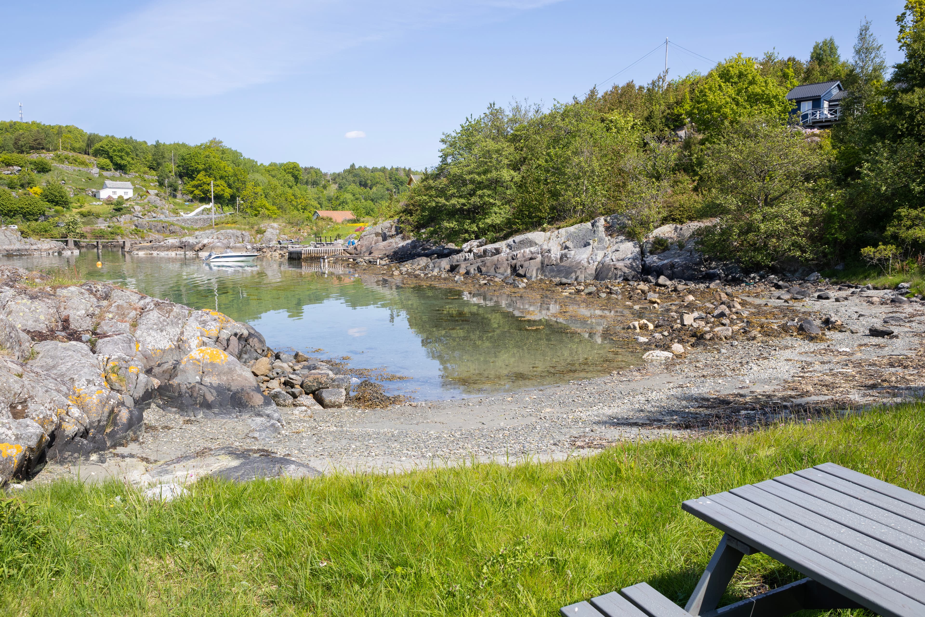 Badestrand med steiner, tare, benk, gress, hytte, liten trekai og blå himmel