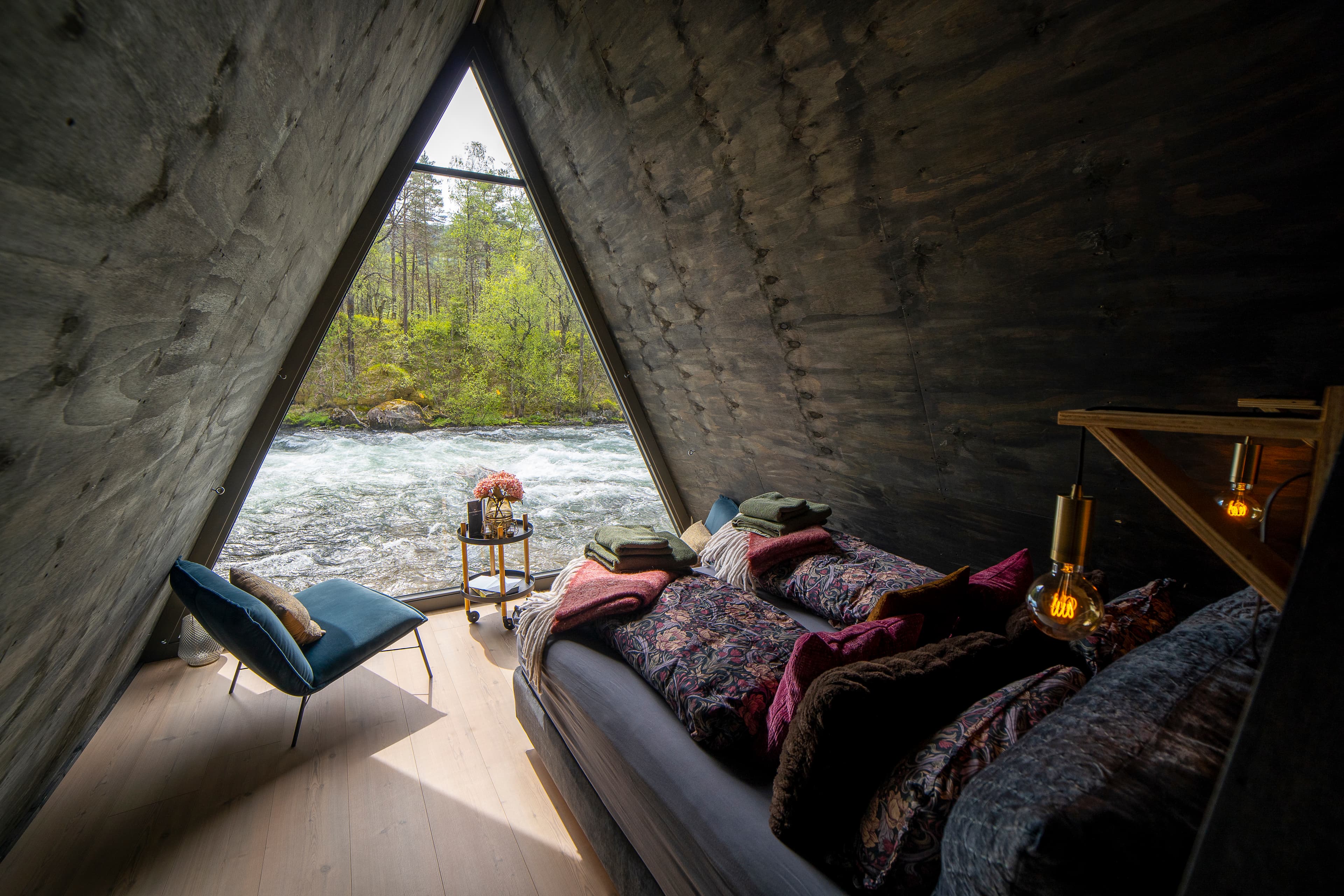 Inne frå ei av hyttene. Hytta er bygd som ein omvendt V og vindauget går frå tak til golv. Det står to blå stolar i front av vindauget, saman med eit lite bord på hjul der det står ein vase med blomar. Senga er i mørke blåfargar og har puter i lilla og brunt. Sengetøyet er mønstra i blå/lilla/gule farger. På enden av senga ligg eit pledd og to handdukar til kvar person (rom for to). Ved hovudenden av senga heng det ei par lamper.