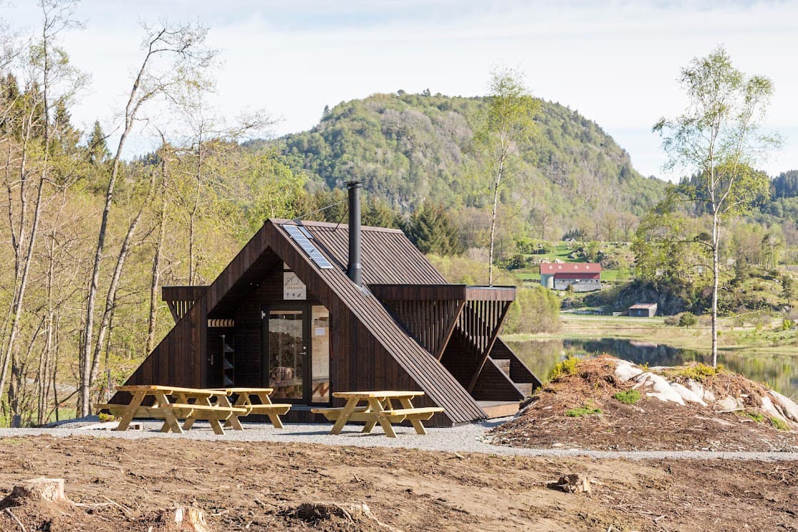 Hytte med benker utenfor, ligger ved vann, bjørk og fjell i bakgrunnen.