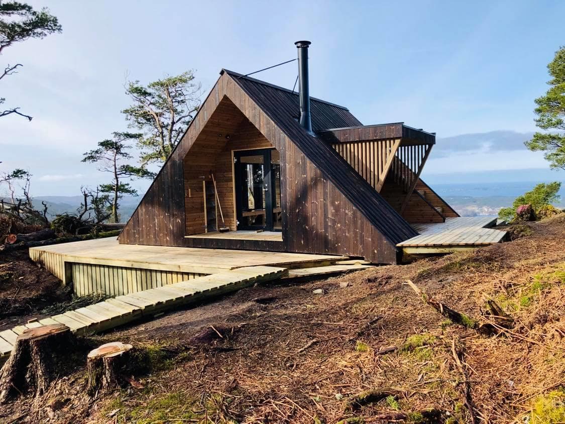 Hytte med terrasse, furutrær og skogbunn.