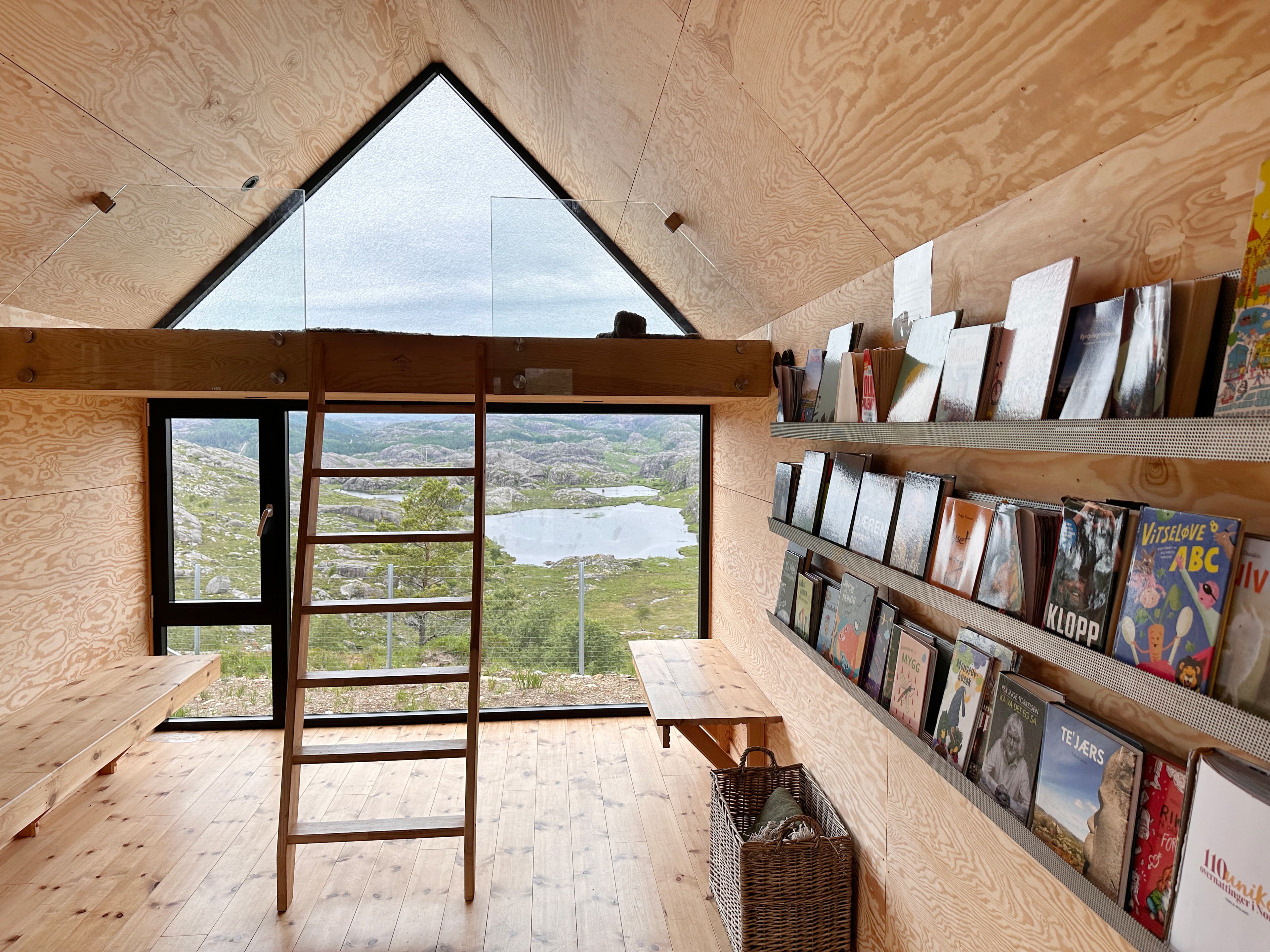 The inside of a cabin with a view of the mountains and lakes. The wooden interior shows a bookshelf with books on the right side, two benches and a small mezzanin with a ladder.