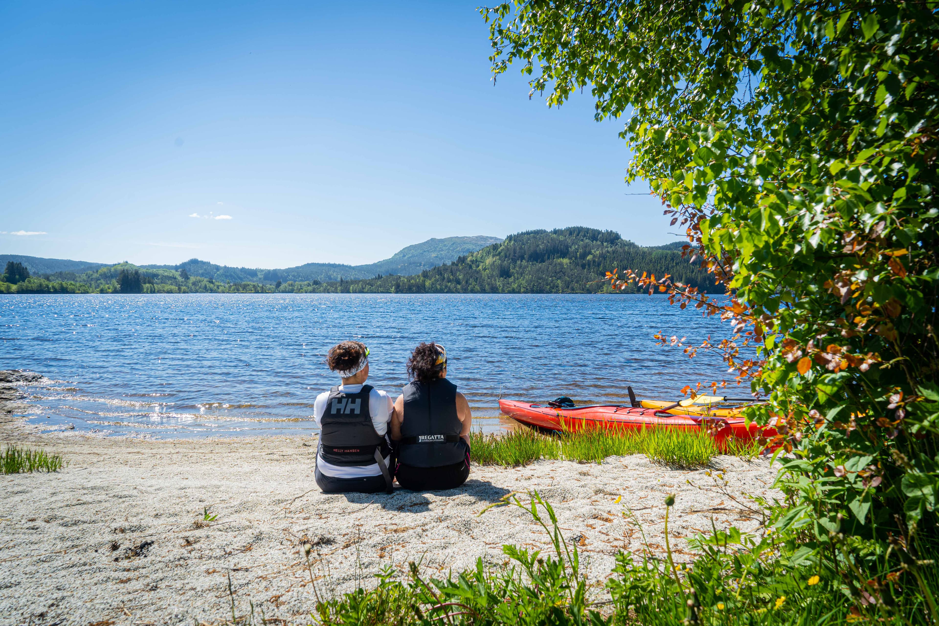 to damer ser ut over vatnet. sitter på ein strand. ved siden av dei ligg det to kajakker. Foto