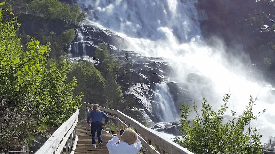 To personar i ei tretrapp ved sida av fossen Langfoss. Mykje vatn i fossen, sol. Foto.