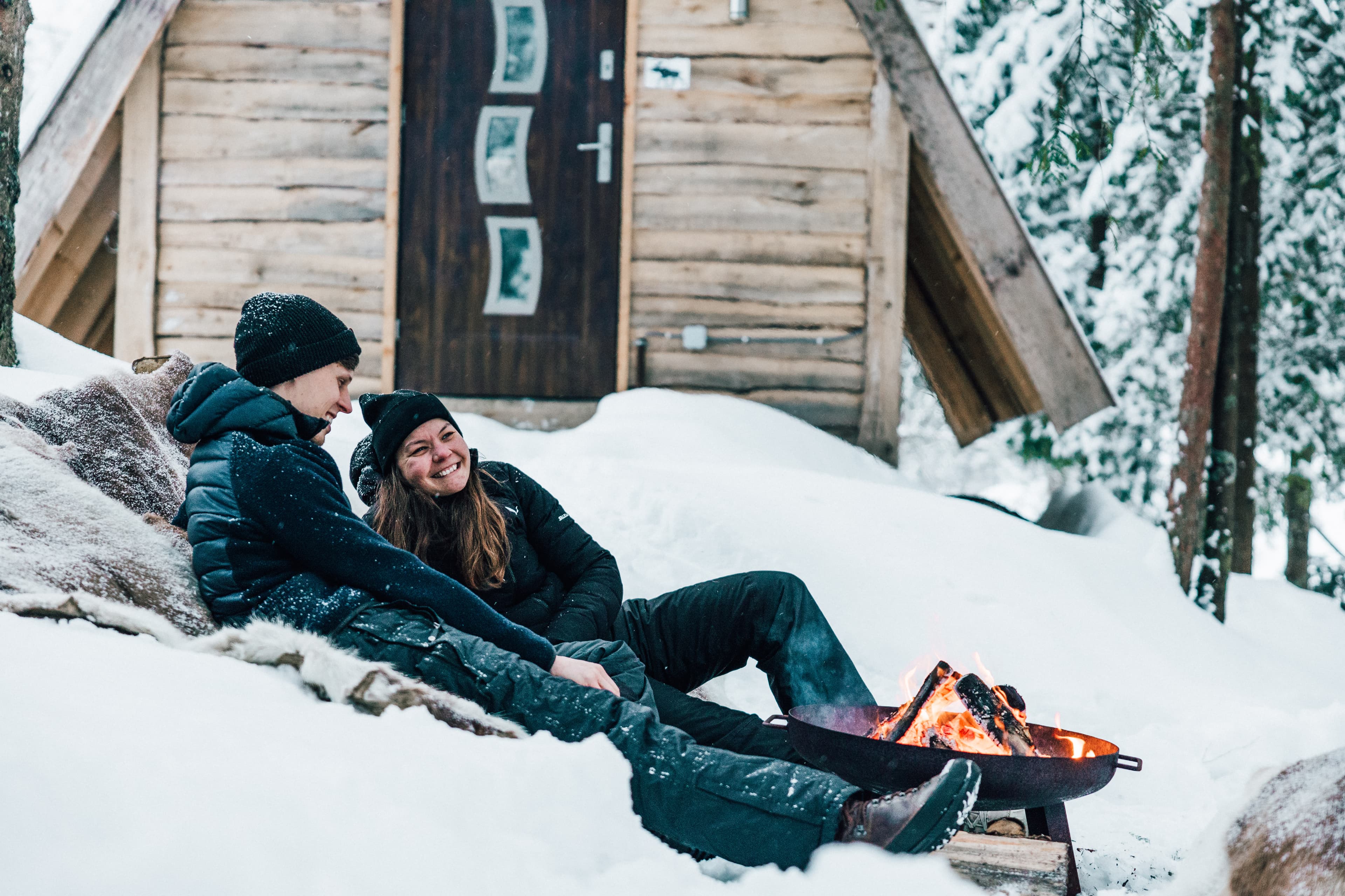 Tee-pee cabins in the snow in Sirdal. A couple in front of the cabin lying in front of a fire smiling