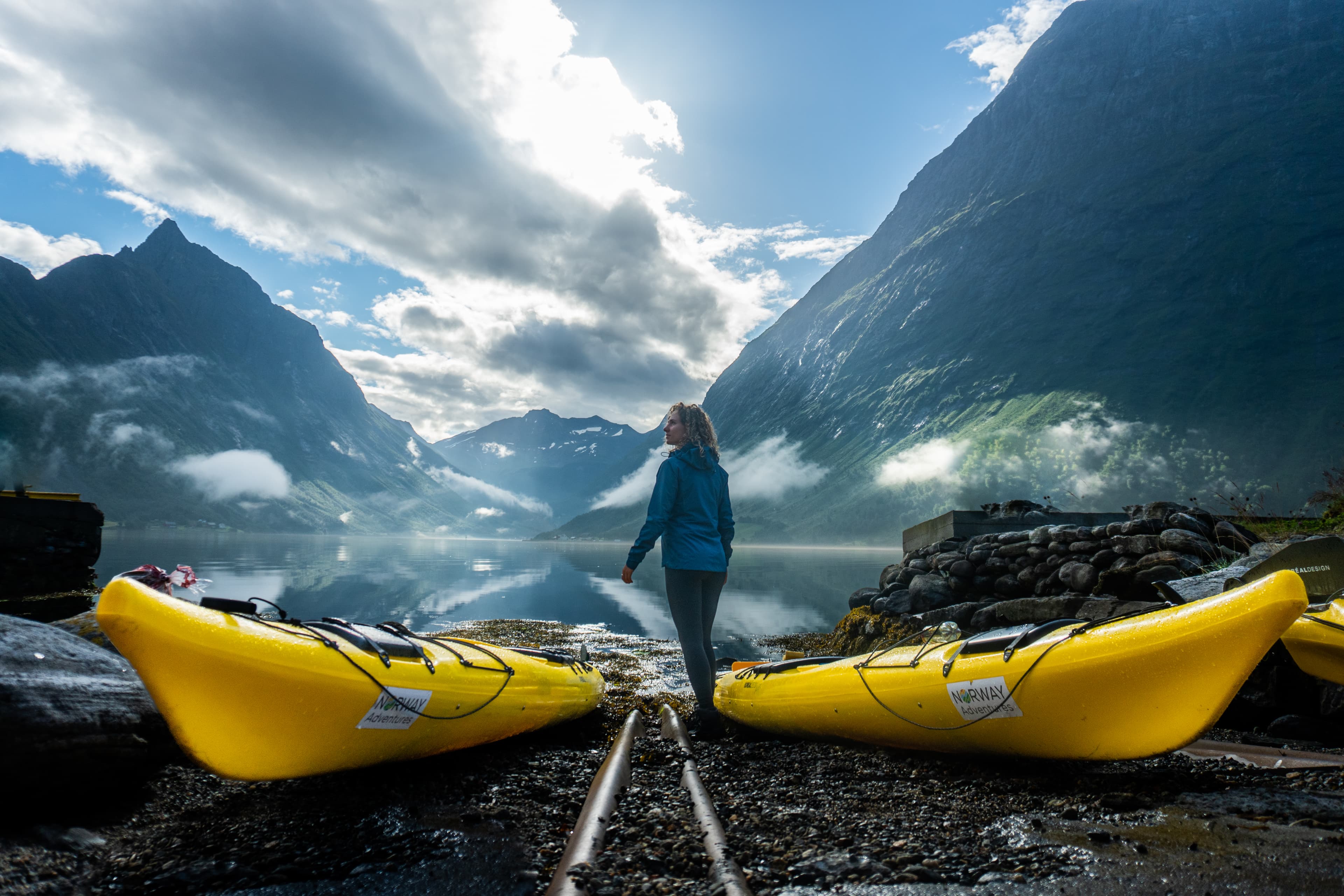 Person med to kajakker står på strand i Hjørundfjorden med fjell i bakgrunnen