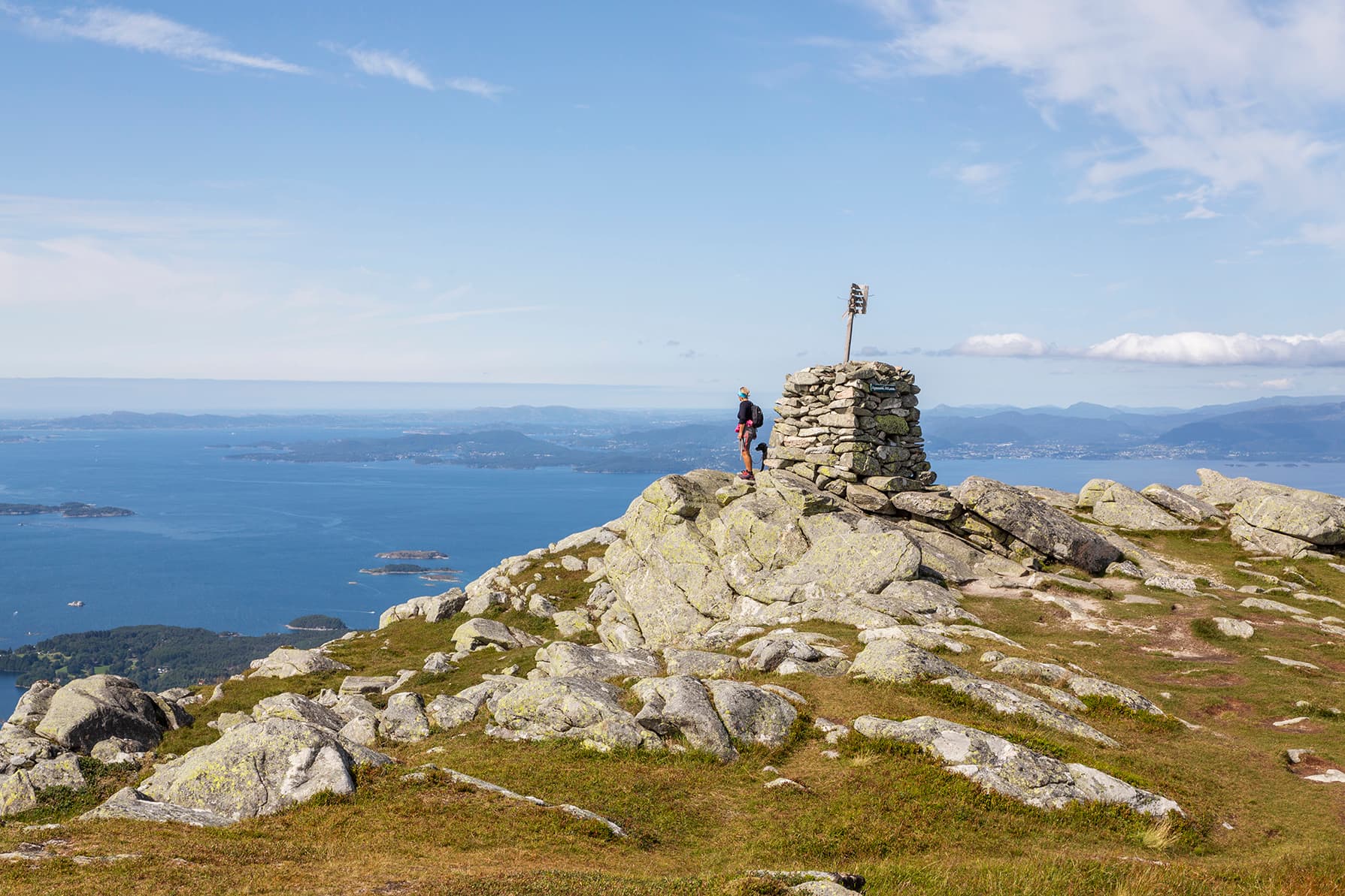 Person og hund som står ved varde på fjelltopp med utstikt mot fjord, fjell og horisonten.