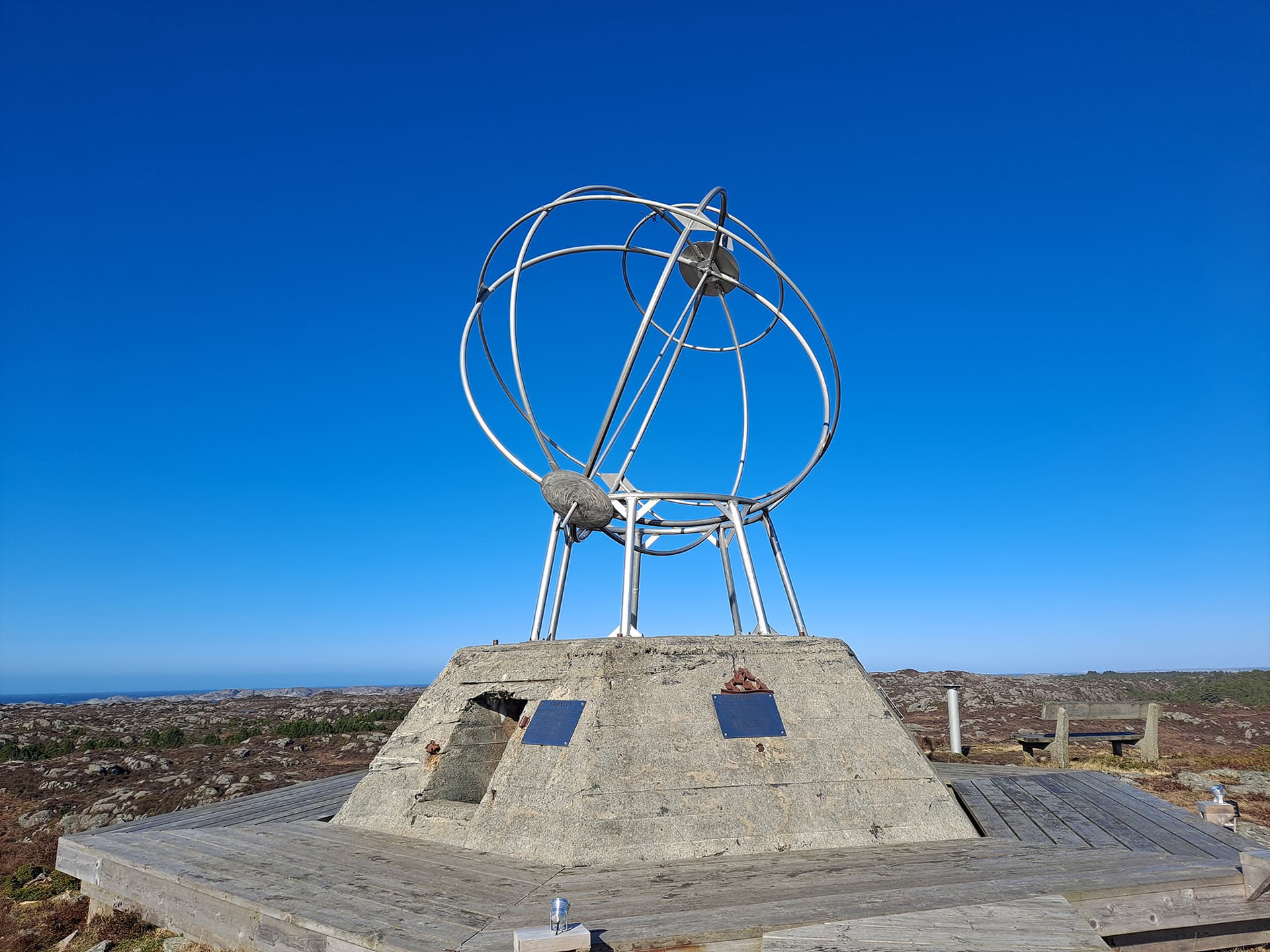 Monument som liknar ei kule på sokkel av mur. Kystlandskap og blå himmel. Foto.