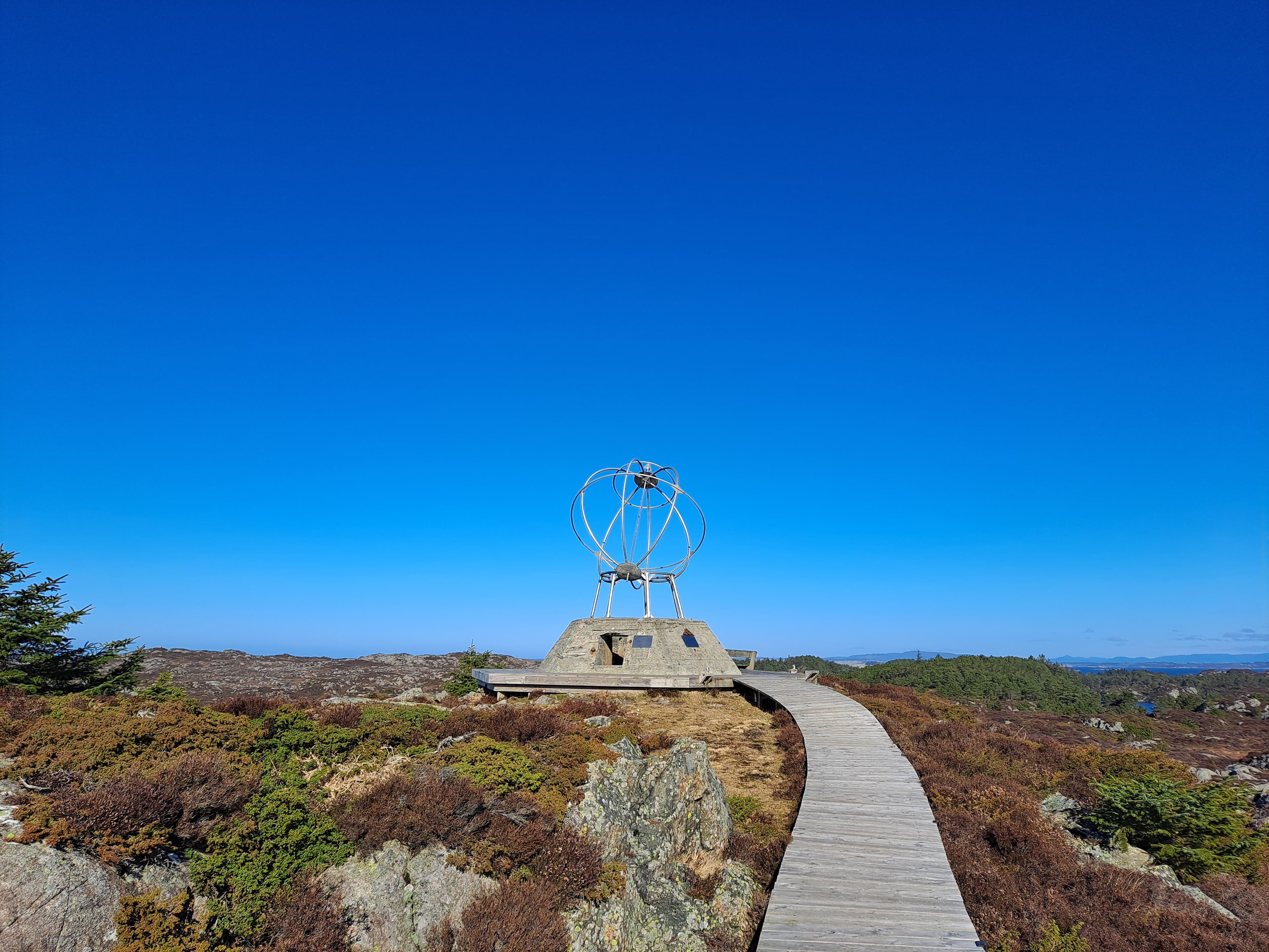 Sti belagt med tettskrudde planker, disse leder til et monument som ligner jordkloden, utsyn mot skog, kystlandskap og hav.