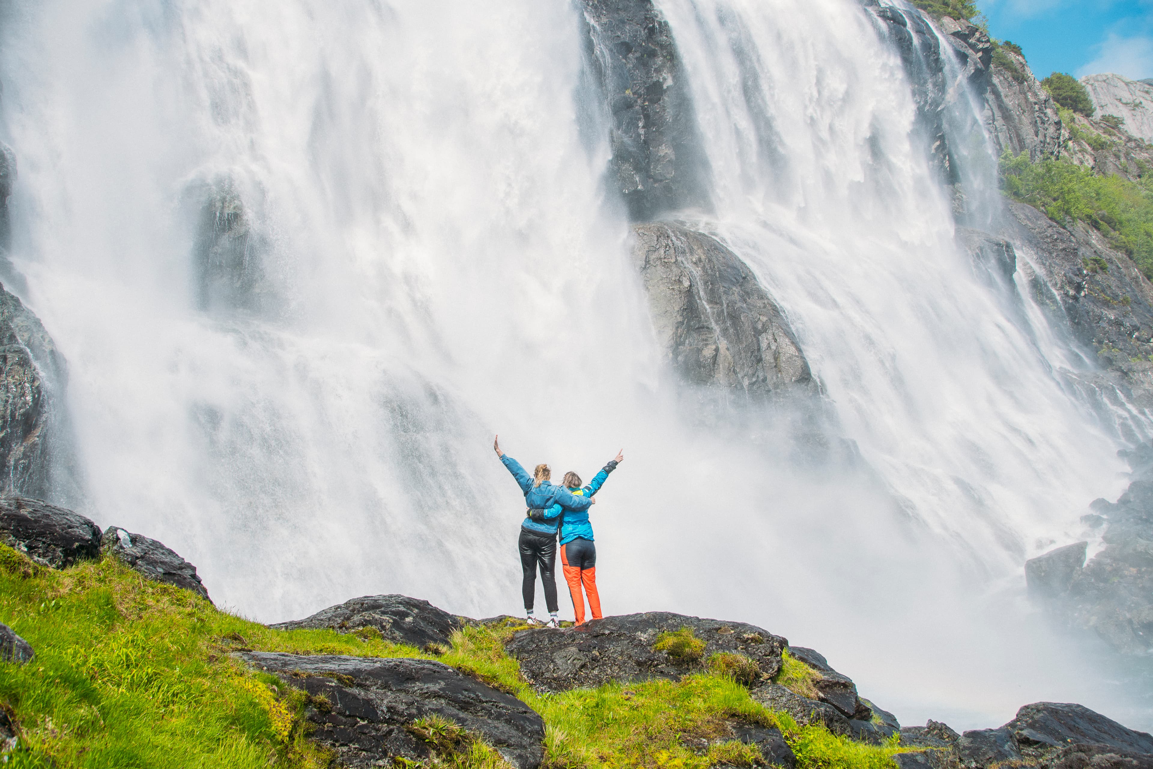 To personar står framfor fossen. Dei held kring kvarandre og held hendene i været. Dei står med ryggen til fotografen. Foto