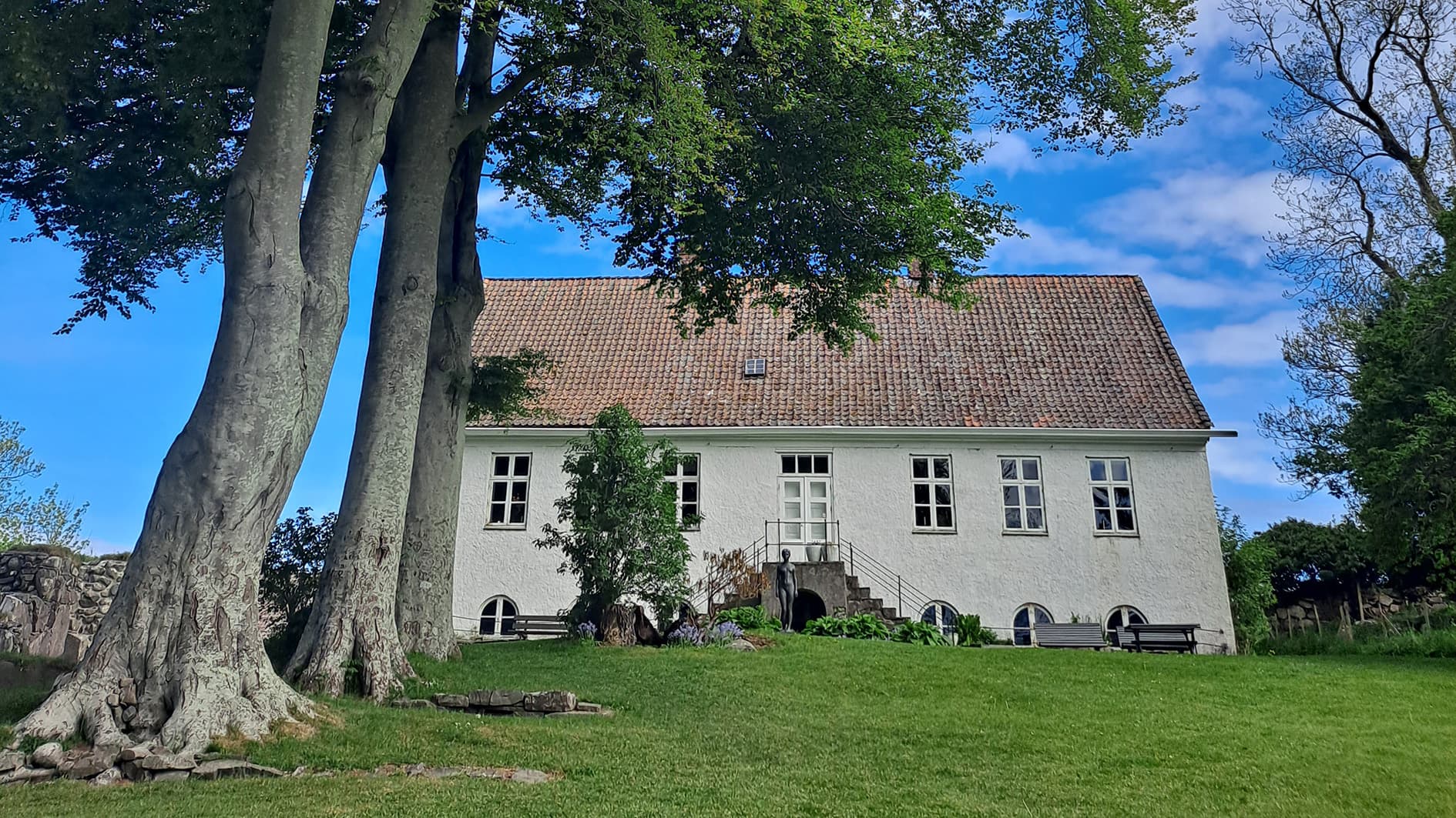 Gamal bygning i mur, raudt tak. trapper utanfor, grøn bakke og store lauvtre. Foto.