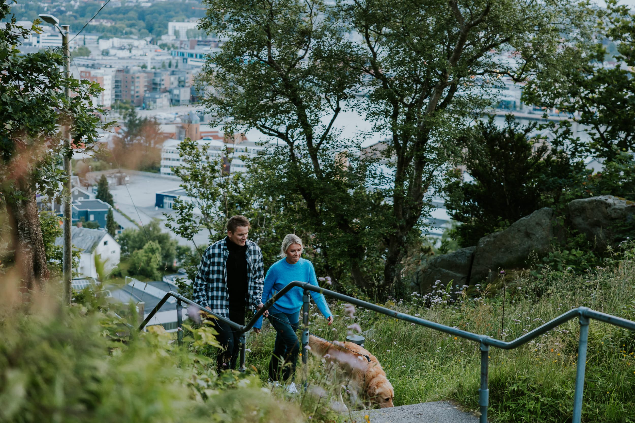 Two people and a dog walking up the stairs to the mountain Hanafjellet. The stairs are called Hanatrappene. From the top the view shows Sandnes and fjord