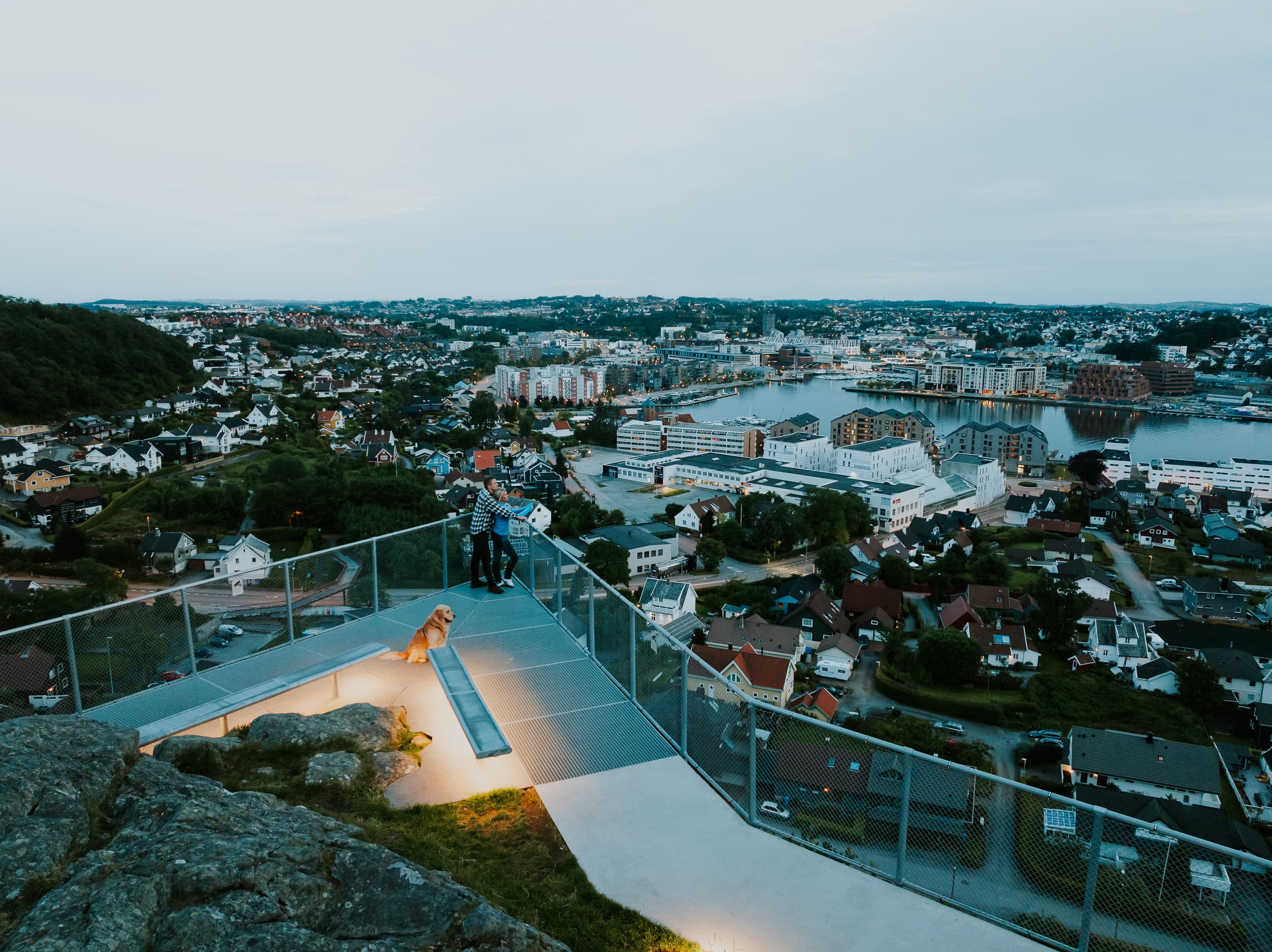 Two people and a dog walking up the stairs to the mountain Hanafjellet. The stairs are called Hanatrappene. From the top the view shows Sandnes and fjord