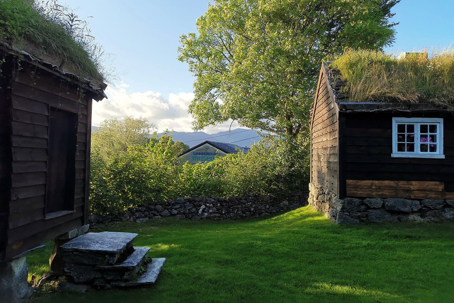 Foto av to eldre hus, steinmur, grønn bakke, stort lauvtre, blå himmel.