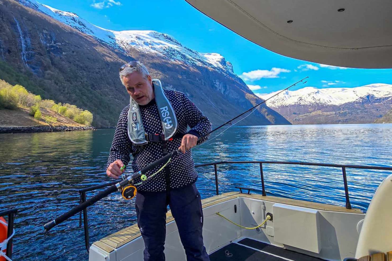 Guiden står i båten og ordner fiskestanga, med geirangerfjorden og fjellene i bakgrunnen.