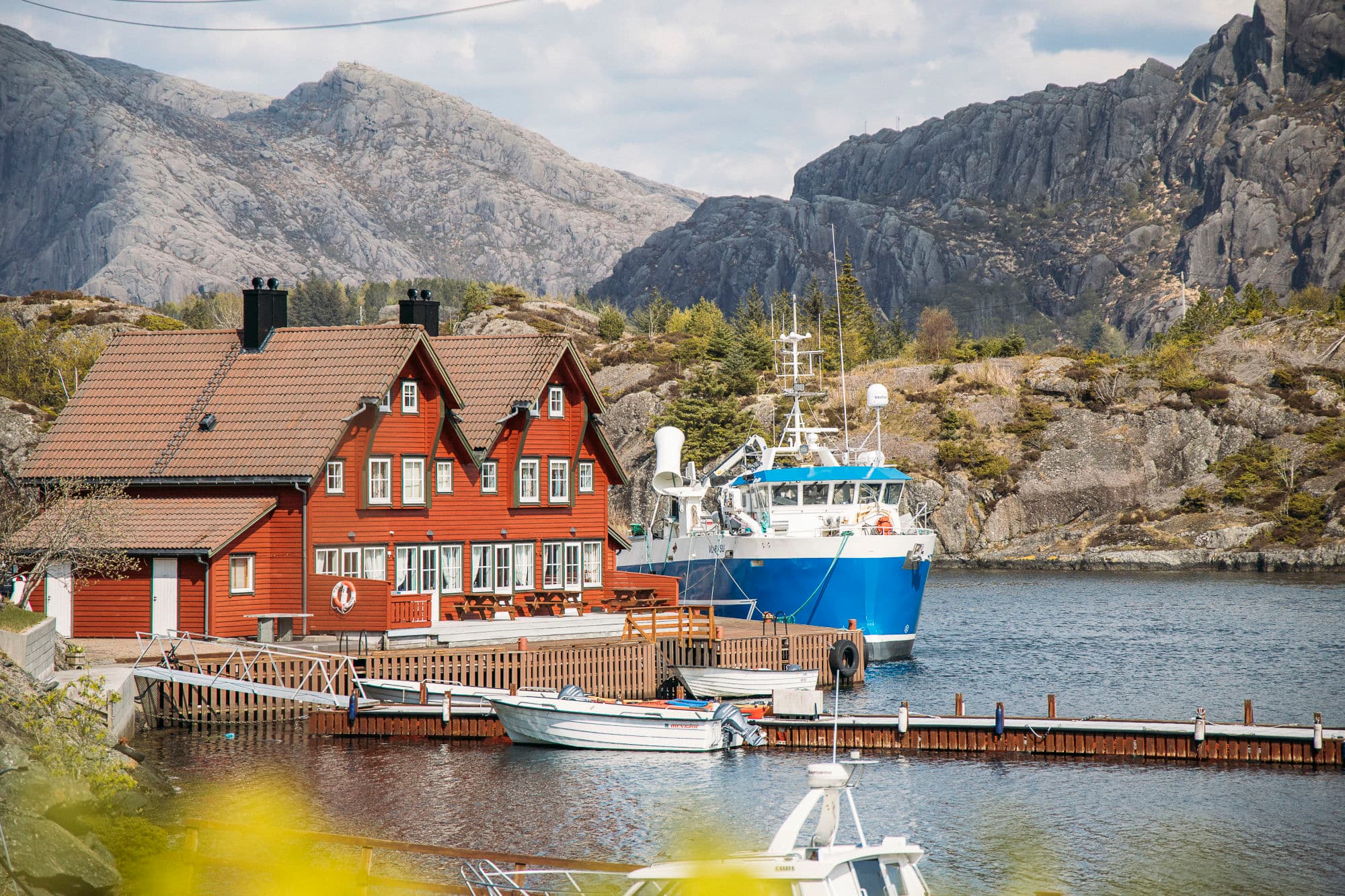 mørke skyer, rorbuer med kai foran og to båter som ligger ved kai. det er fjell i bakgrunnen. fiskebåt ligger ved siden av marinesenteret