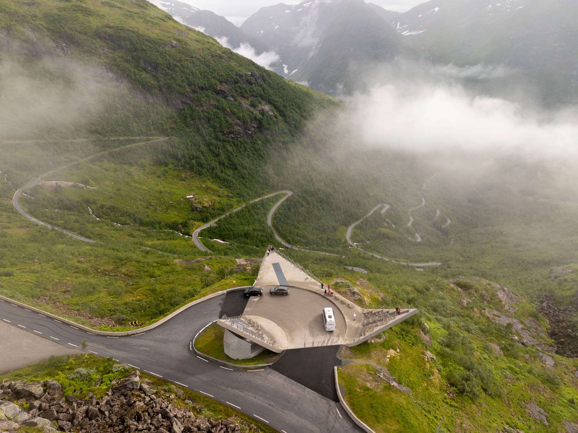 utsiktspunkt med veg nedenfor. fjell, grønne trer og en firkanta utsiktspunkt. tre biler står der