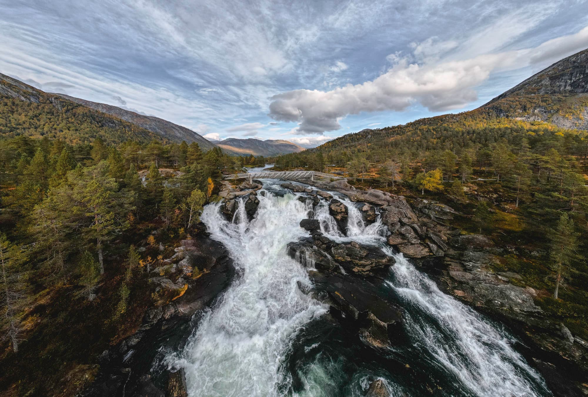 en foss som går langs en sti. det er oransj skog rundt fossen. høge fjell sees i horisonten. det går en bro over fossen