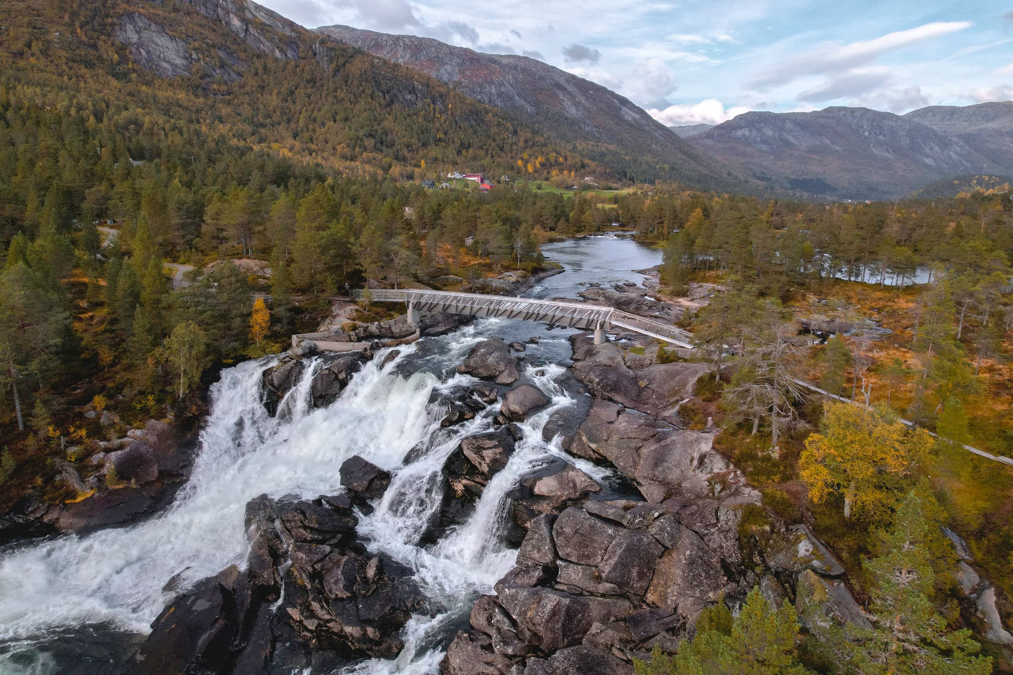 en foss som går langs en sti. det er oransj skog rundt fossen. høge fjell sees i horisonten. det går en bro over fossen