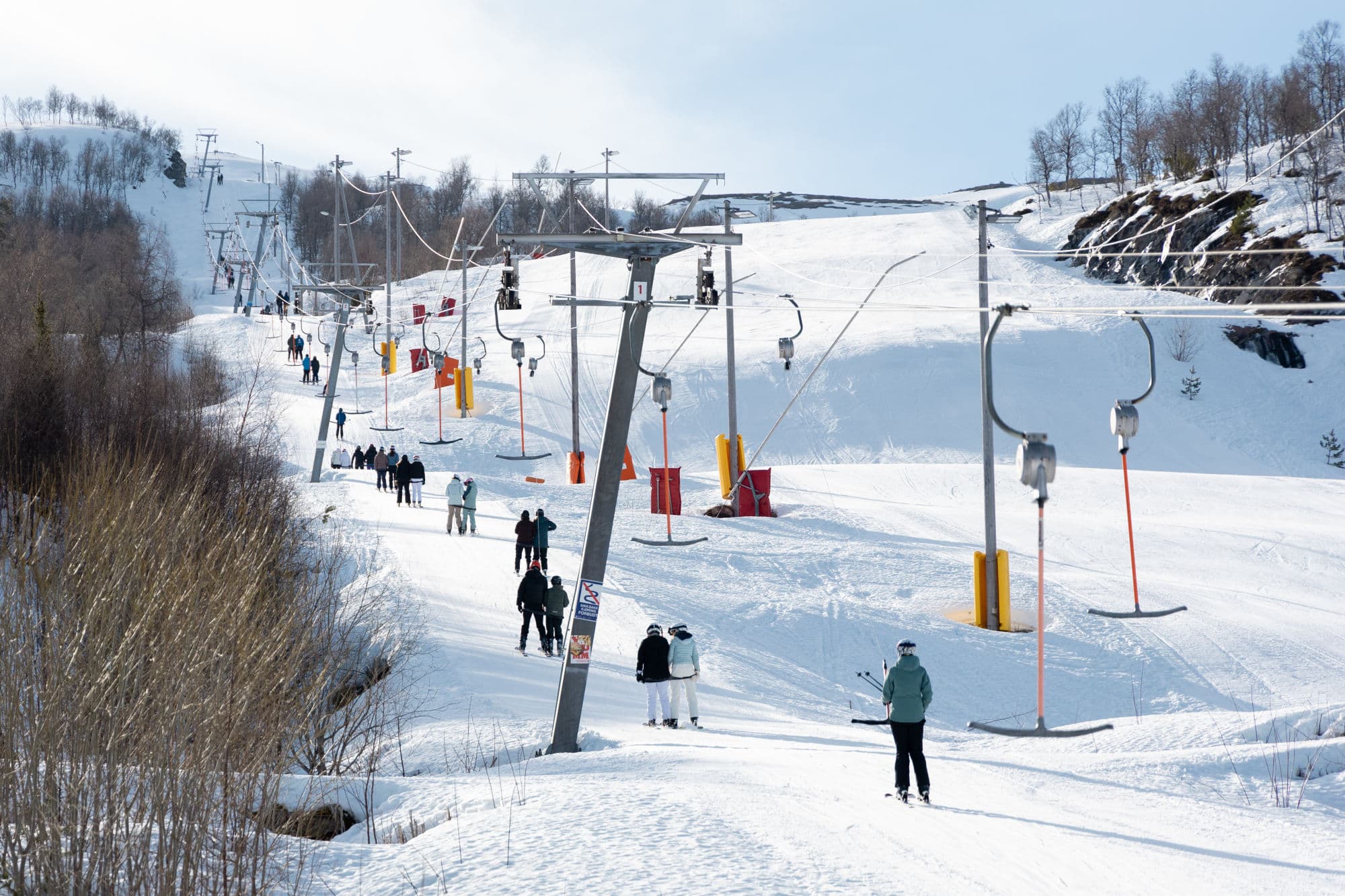 mange personer står i eit t-trekk opp en skibakke. det er sol og blå himmel og snø på bakken